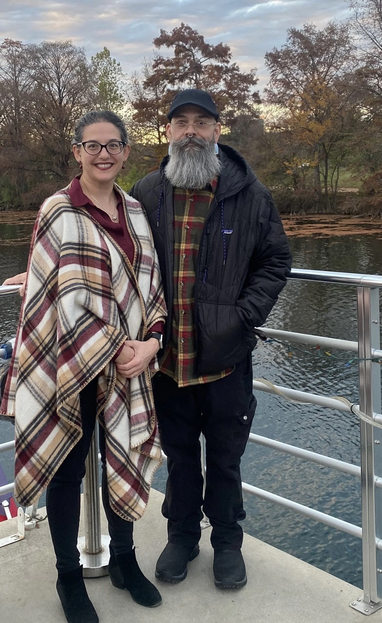 A smiling woman and a man with a gray beard standing on a boat or dock near a body of water with trees in the background during autumn.