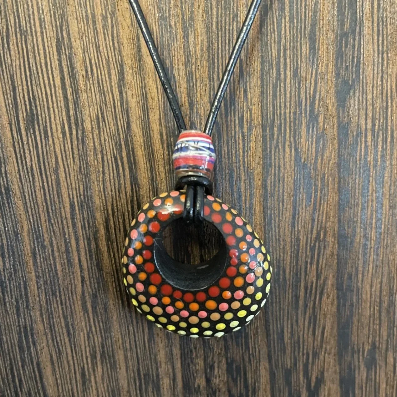 This one-of-a-kind pendant necklace features a natural stone with a hand-drilled hole, transformed into wearable art through the intricate dot painting technique.