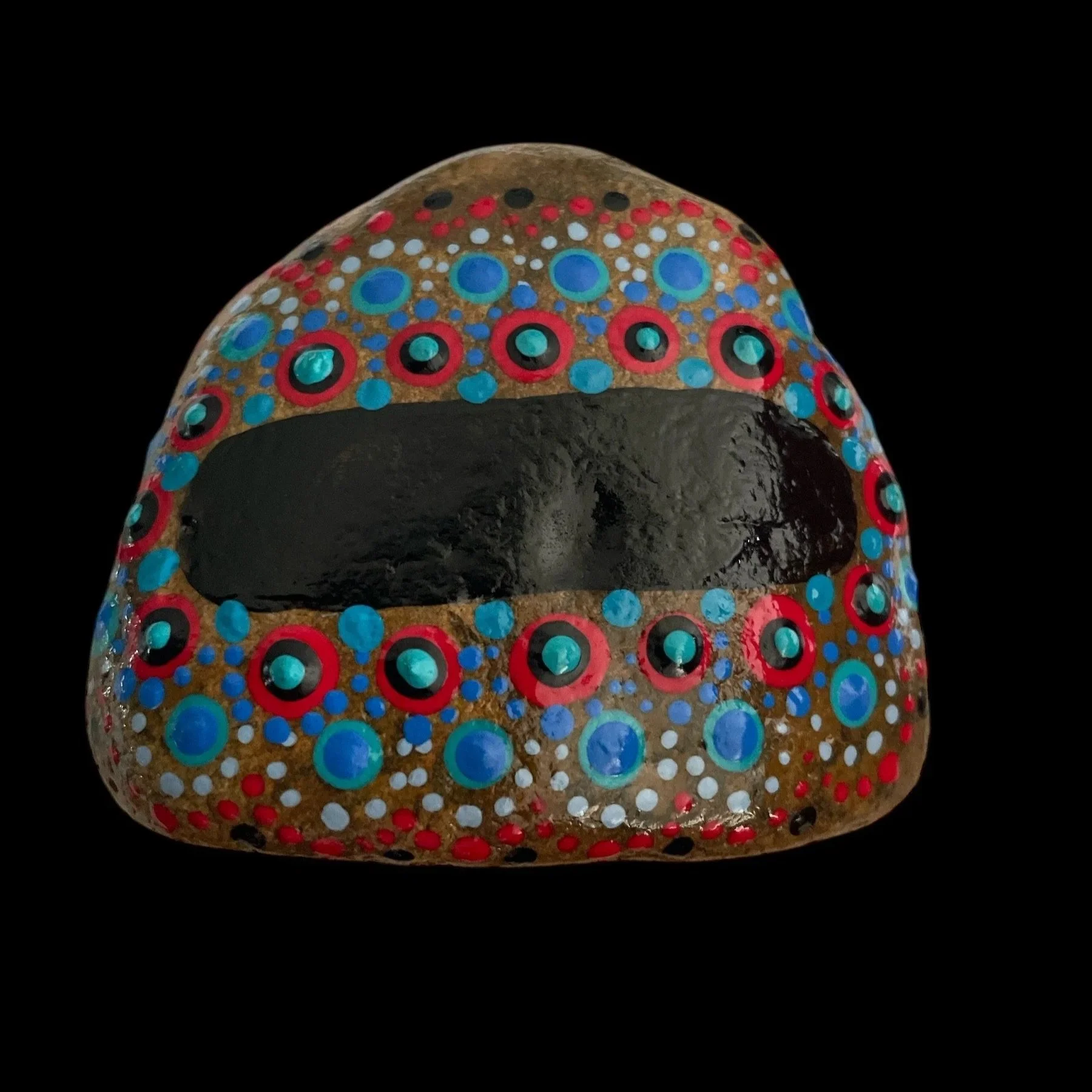 Oval rock with intricate dot painting in concentric circles of red, blue, black, and white on black background