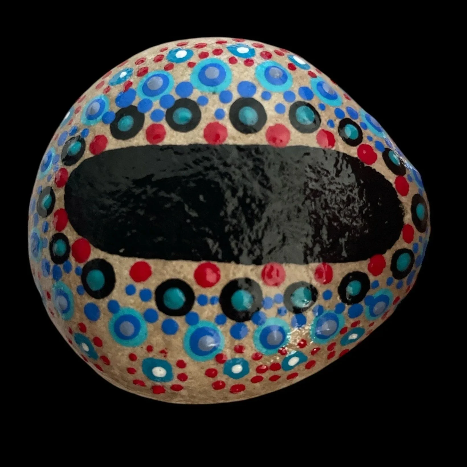 Oval rock with intricate dot painting in concentric circles of red, black, blue, and white on black background