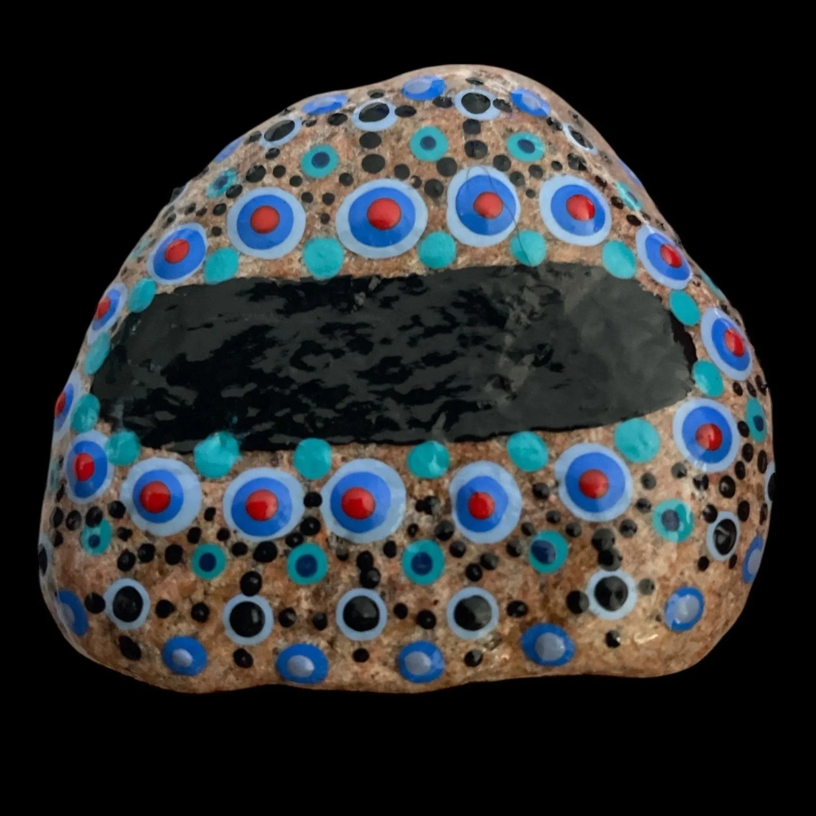 Oval rock painted with concentric circles and dot pattern in blue, black, and red on beige background, featuring central black oval surrounded by decorative rows.