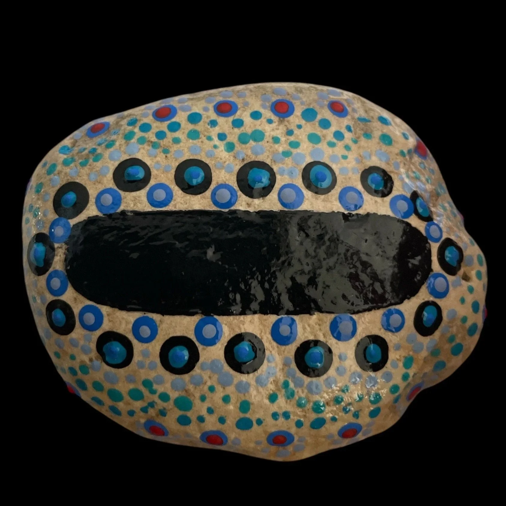 Oval rock painted with concentric circles and dot pattern in blue, black, and red on beige background, featuring central black oval surrounded by decorative rows.
