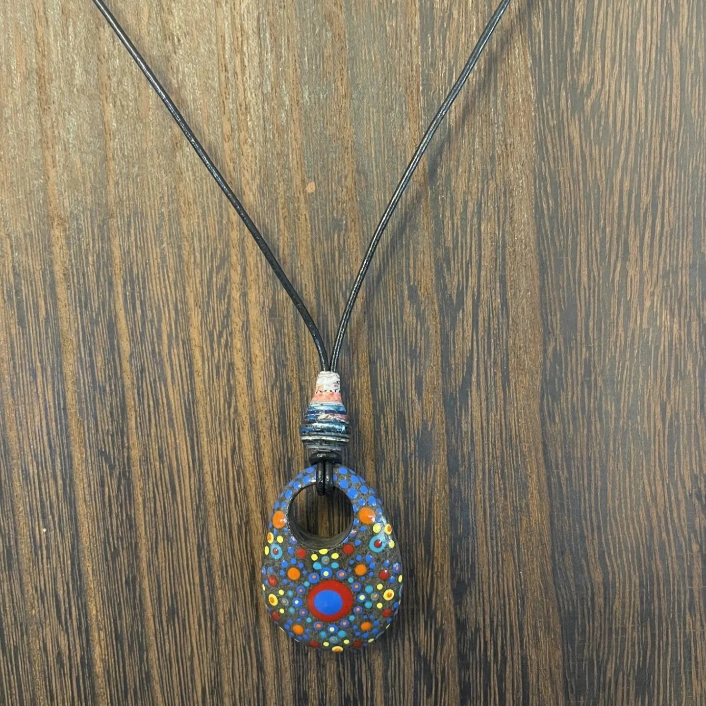 This one-of-a-kind pendant necklace features a natural stone with a hand-drilled hole, transformed into wearable art through the intricate dot painting technique.