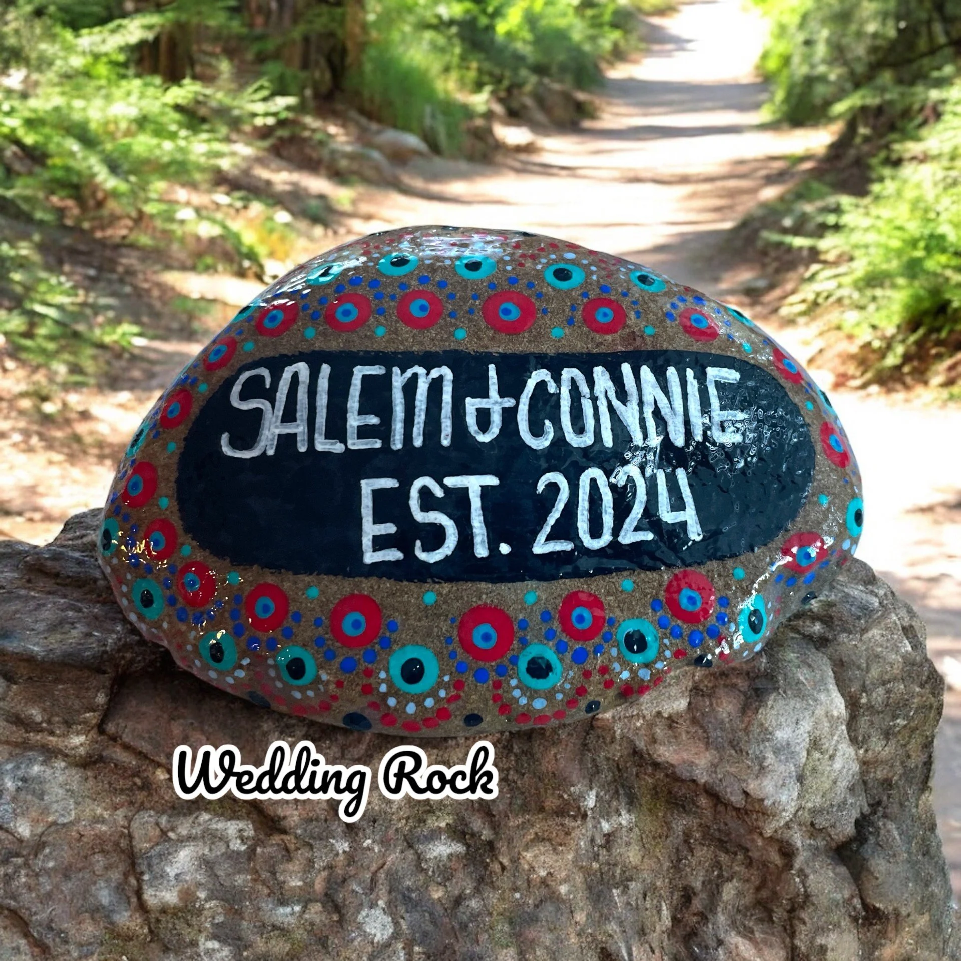 Decorative wedding rock painted with colorful dots, featuring the names Salem and Connie and the year 2024, placed on a rock wall on a dirt trail in a forest setting.