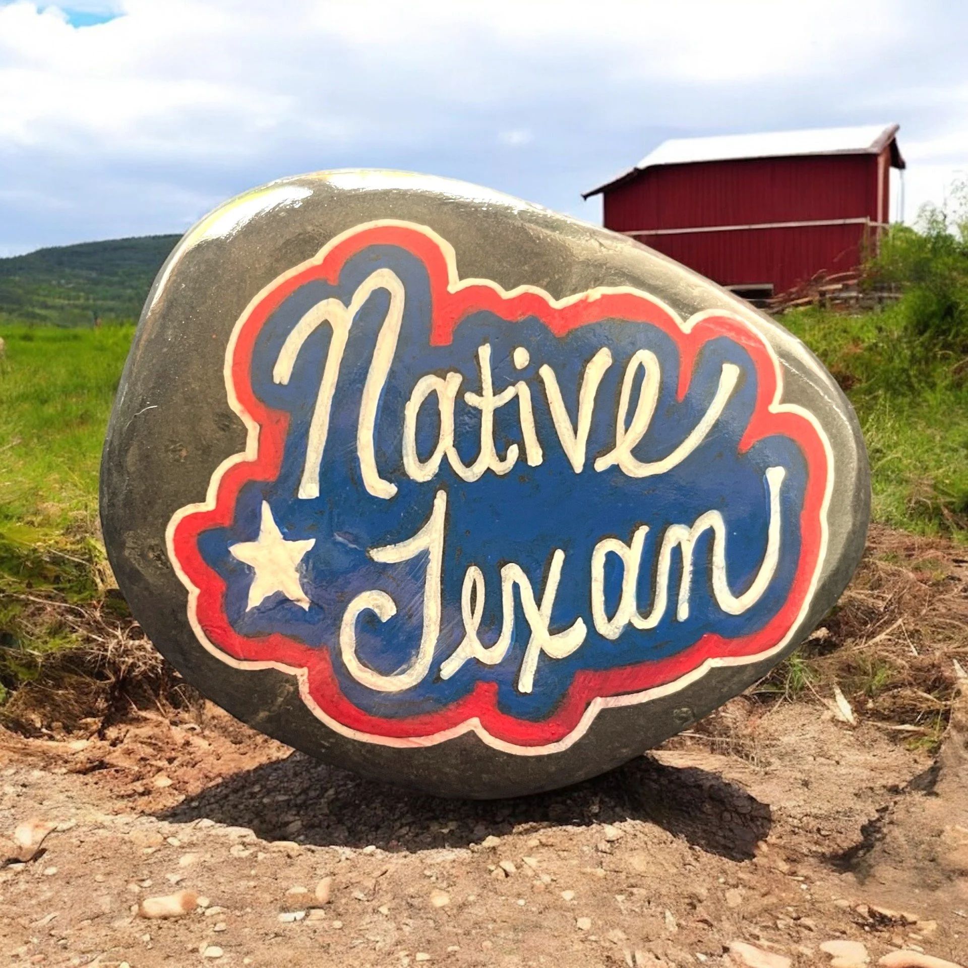 A large stone with the words 'Native Texan' painted in blue and white script, decorated with a star, on a rural farm landscape with a red barn in the background.