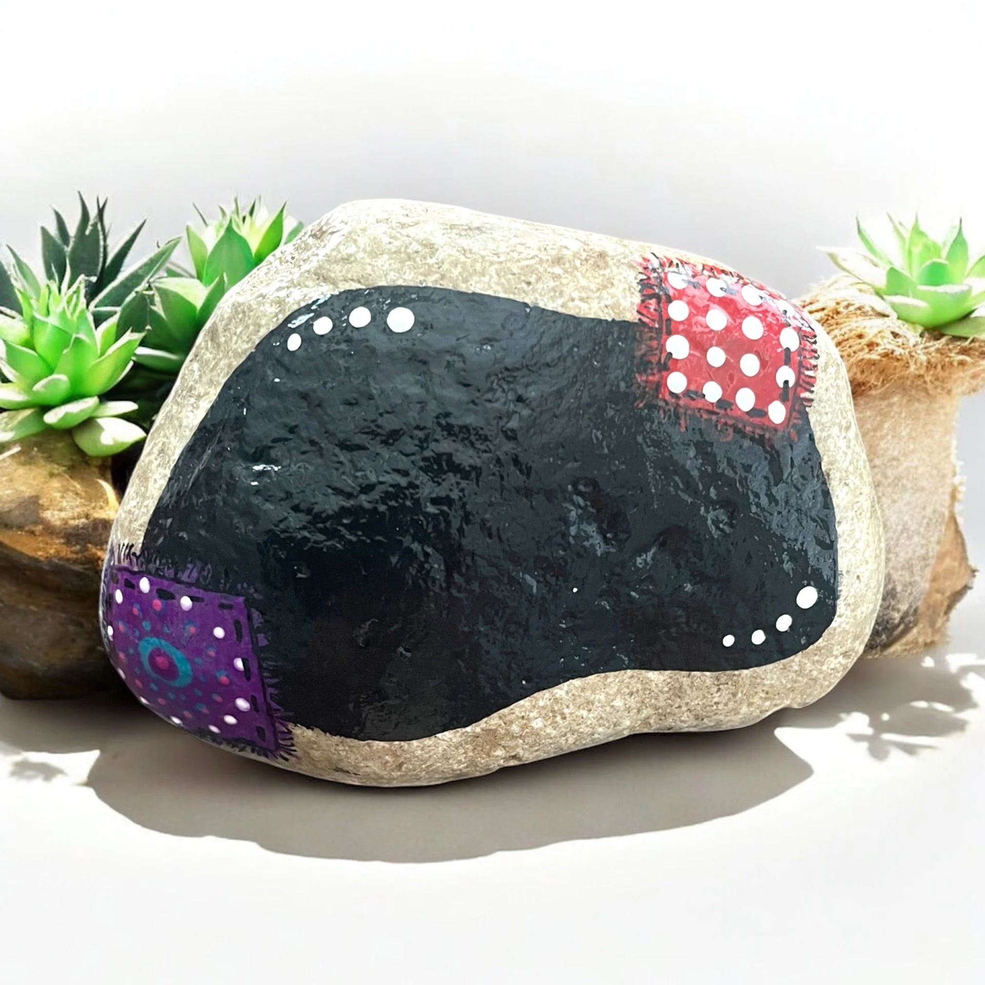 Painted rock of a black chalkboard with a colorful, patterned quilt patches, surrounded by succulents.