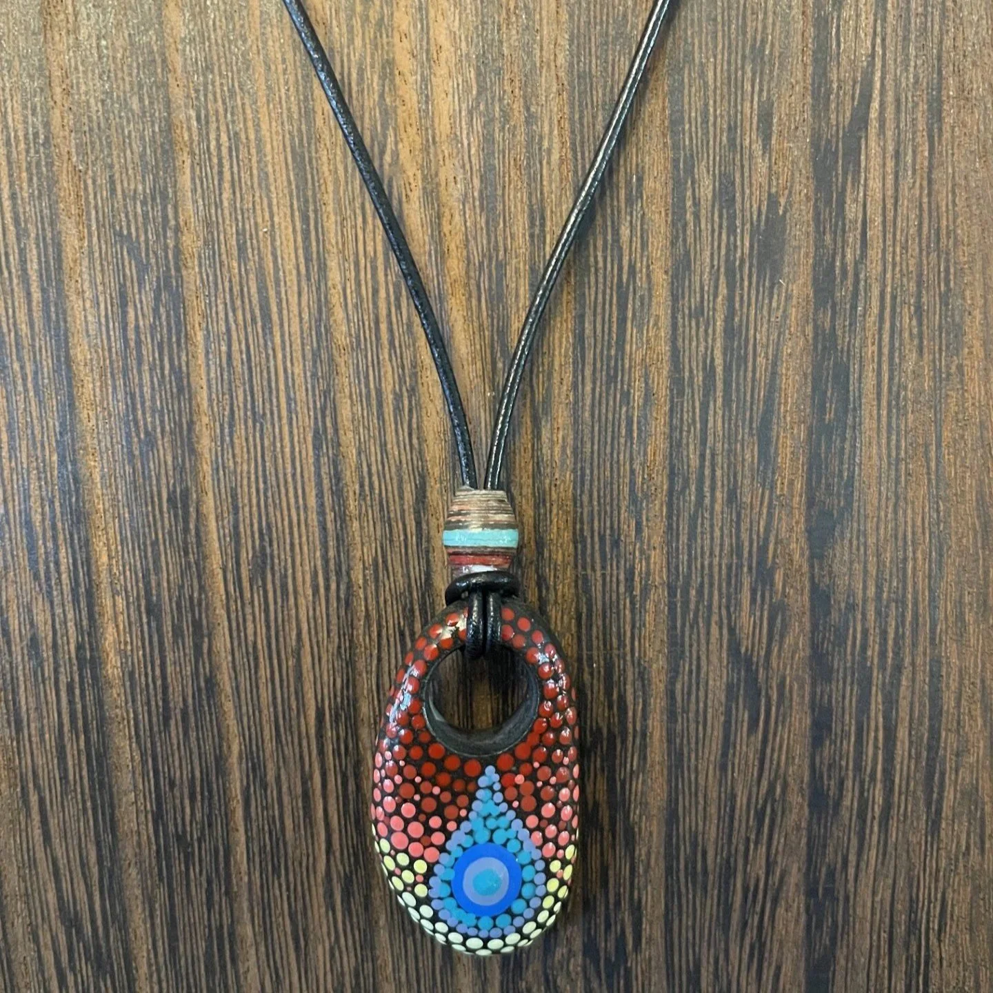 This one-of-a-kind pendant necklace features a natural stone with a hand-drilled hole, transformed into wearable art through the intricate dot painting technique.