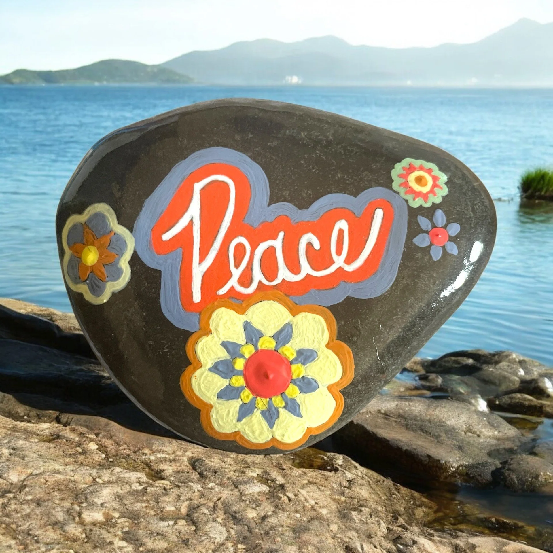 Dark rock painted with 'Peace' and outlined in orange and blue, decorated with colorful floral designs with blue, yellow, and orange.