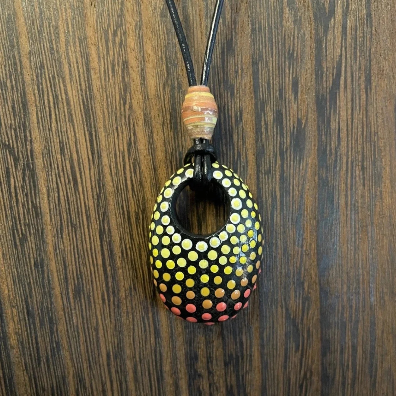 This one-of-a-kind pendant necklace features a natural stone with a hand-drilled hole, transformed into wearable art through the intricate dot painting technique.
