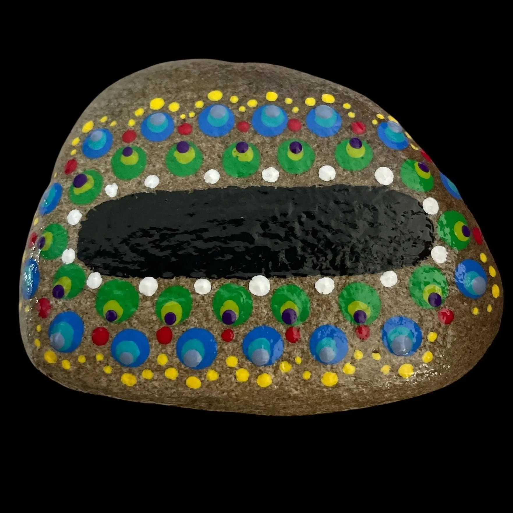 Oval rock painted with colorful dot patterns in concentric circles of blue, green, yellow, red, and white around central black oval on natural stone background.