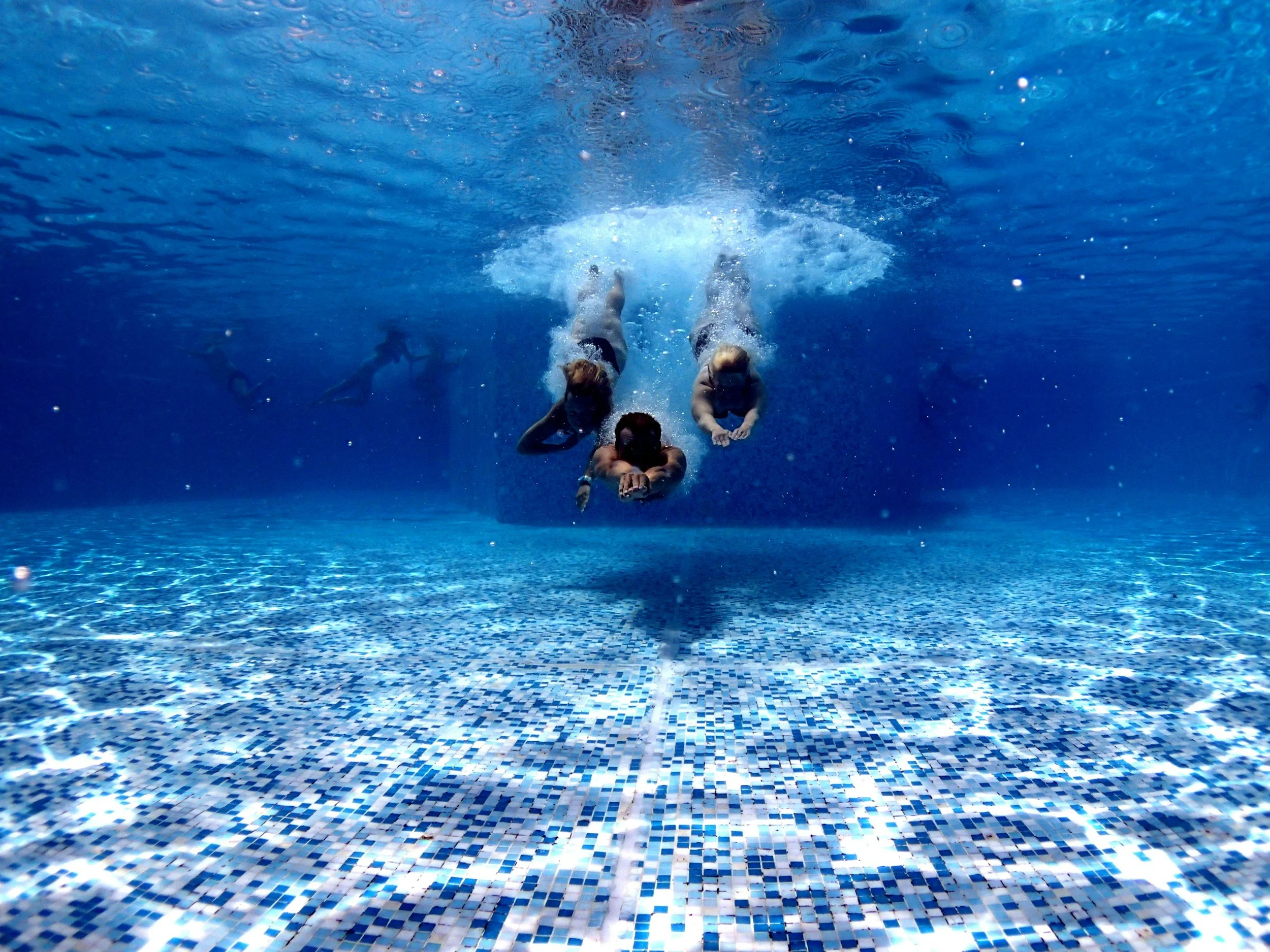 Drei Menschen schwimmen unter Wasser im Swimmingpool, wobei sie nach vorne schauen. Im Hintergrund sind weitere Schwimmer sichtbar.