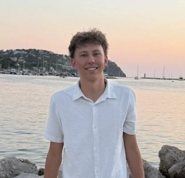 Ein junger Mann steht am Ufer mit Booten und Bergen im Hintergrund bei Sonnenuntergang, trägt ein weißes Kurzarmhemd, lächelt und schaut in die Kamera.