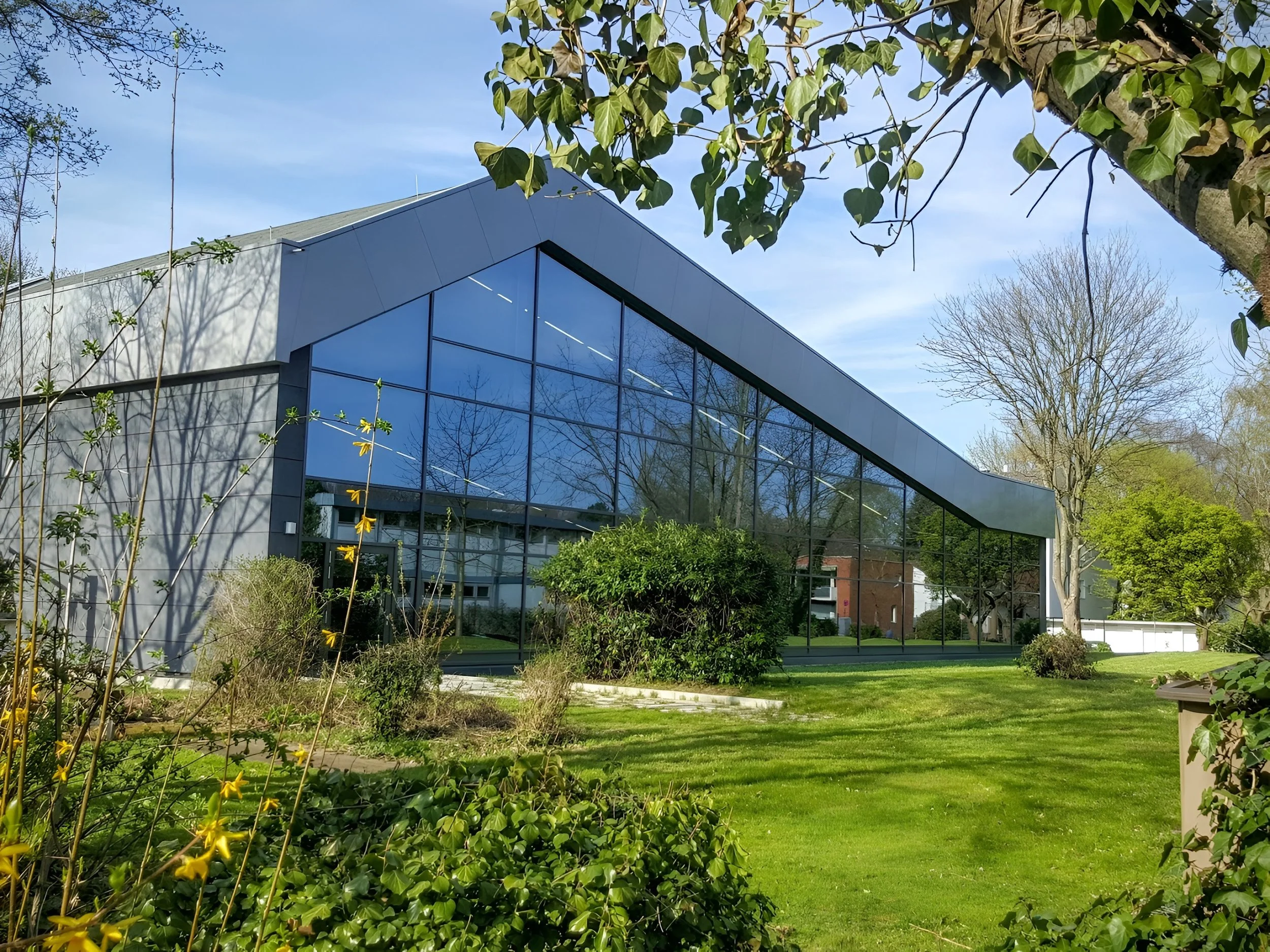 Modernes Gebäude mit großer Glasfassade, umgeben von grünen Bäumen und Rasenflächen, bei schönem Wetter.