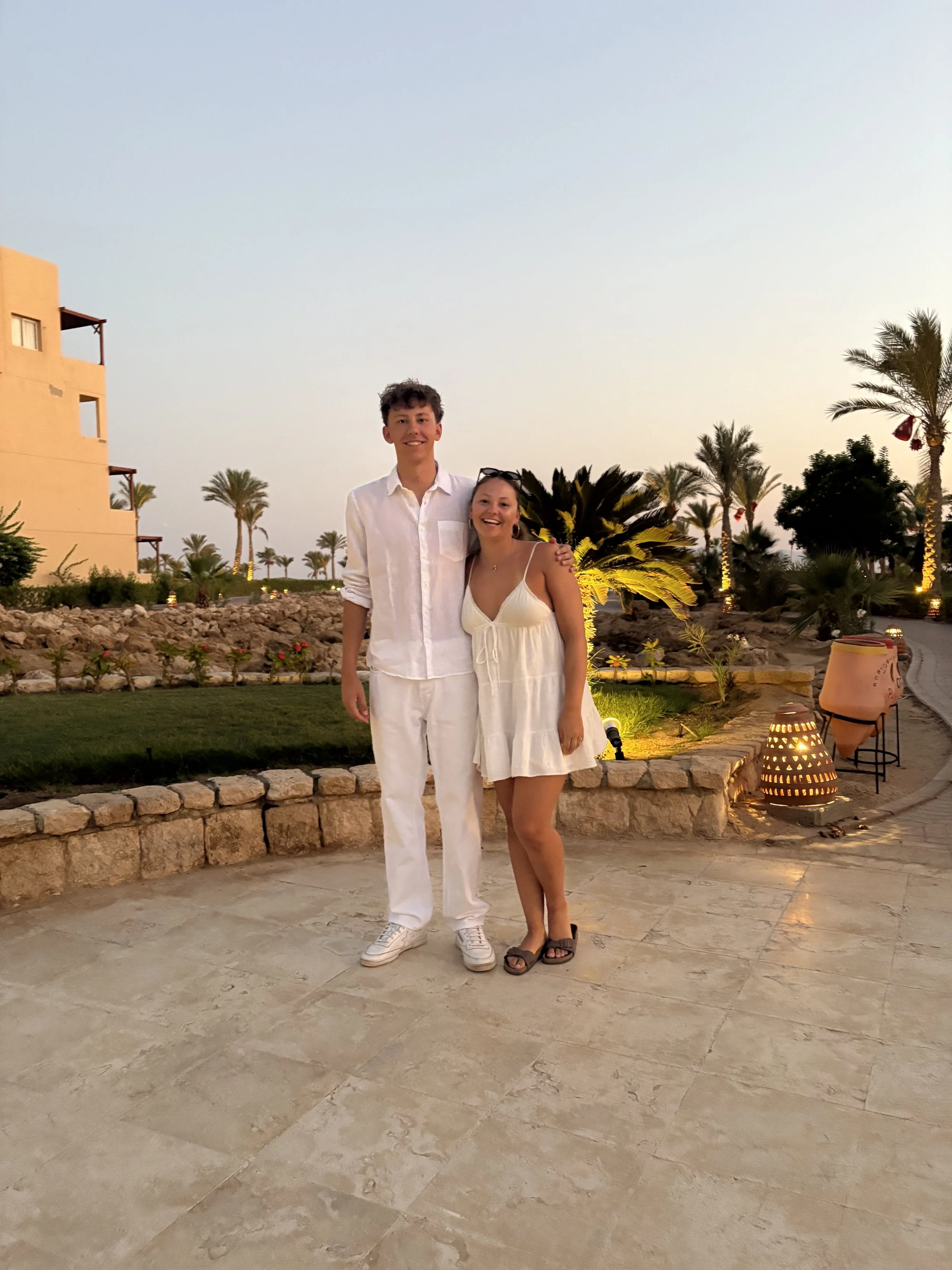 Ein Mann und eine Frau stehen zusammen im Freien bei Sonnenuntergang, lächelnd, umgeben von Palmen und Gartenbeleuchtung, in sommerlicher Kleidung.