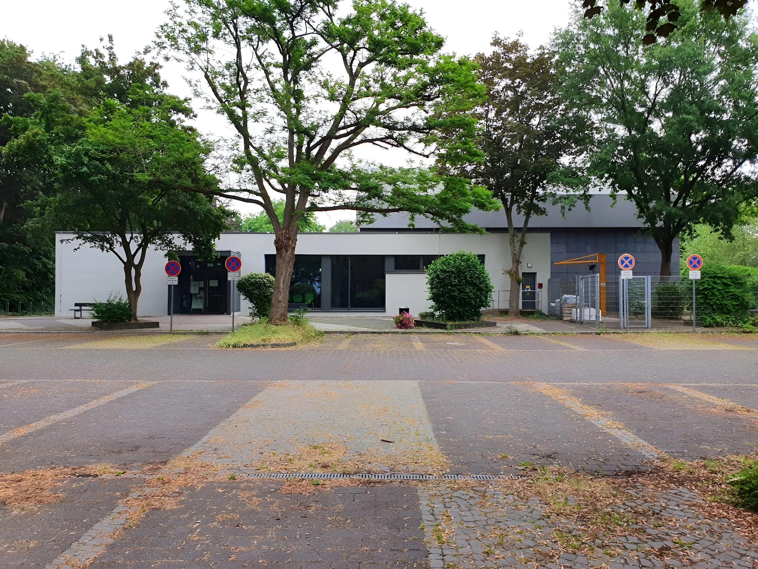 Leeres Parkplatz mit vier Parkverbotsschildern vor einem modernen Gebäude und grünen Bäumen.