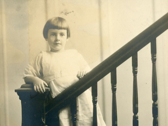 Young girl standing on a staircase in a white dress with her hands on the banister
