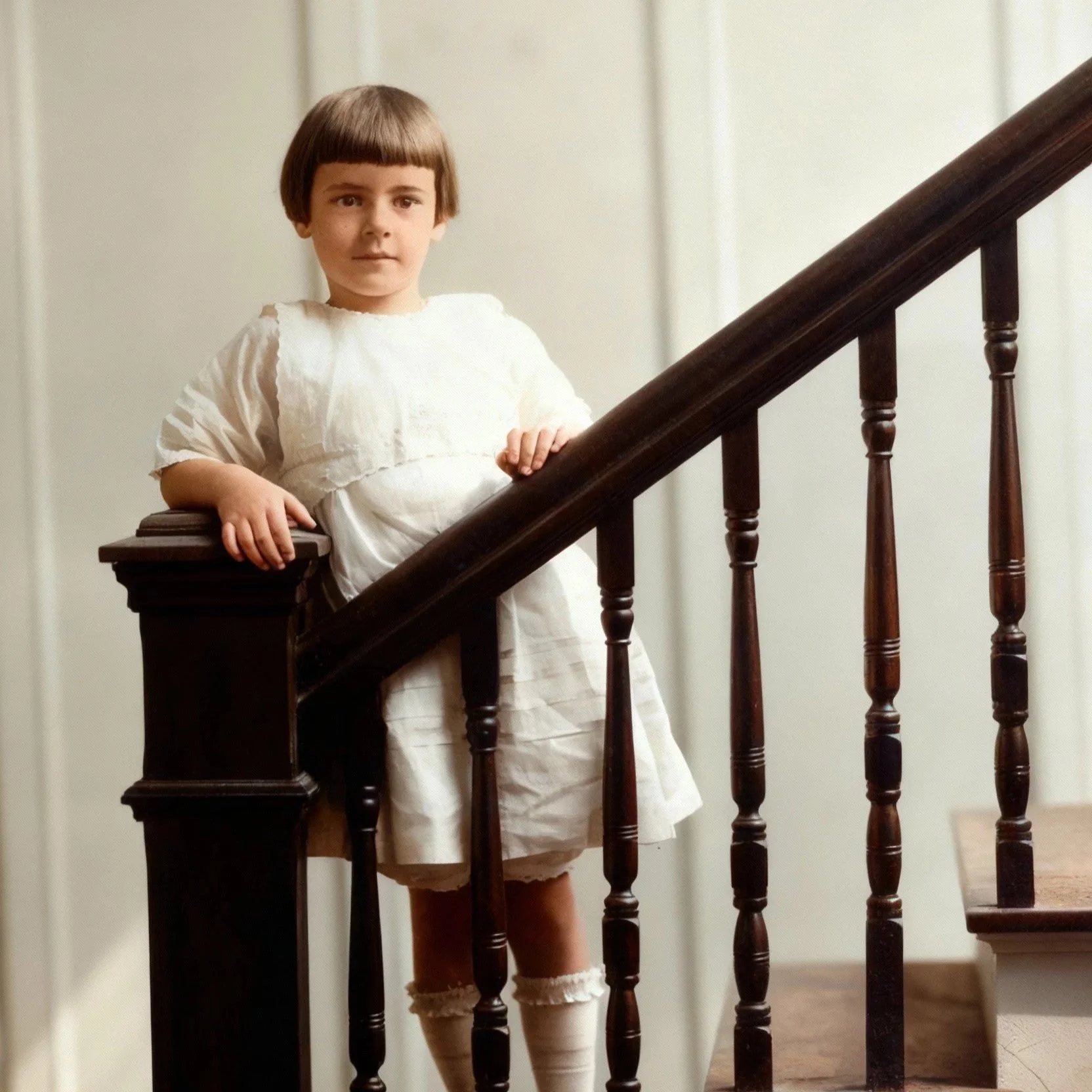 Full color picture of a young girl standing on a staircase in a white dress with her hands on the banister