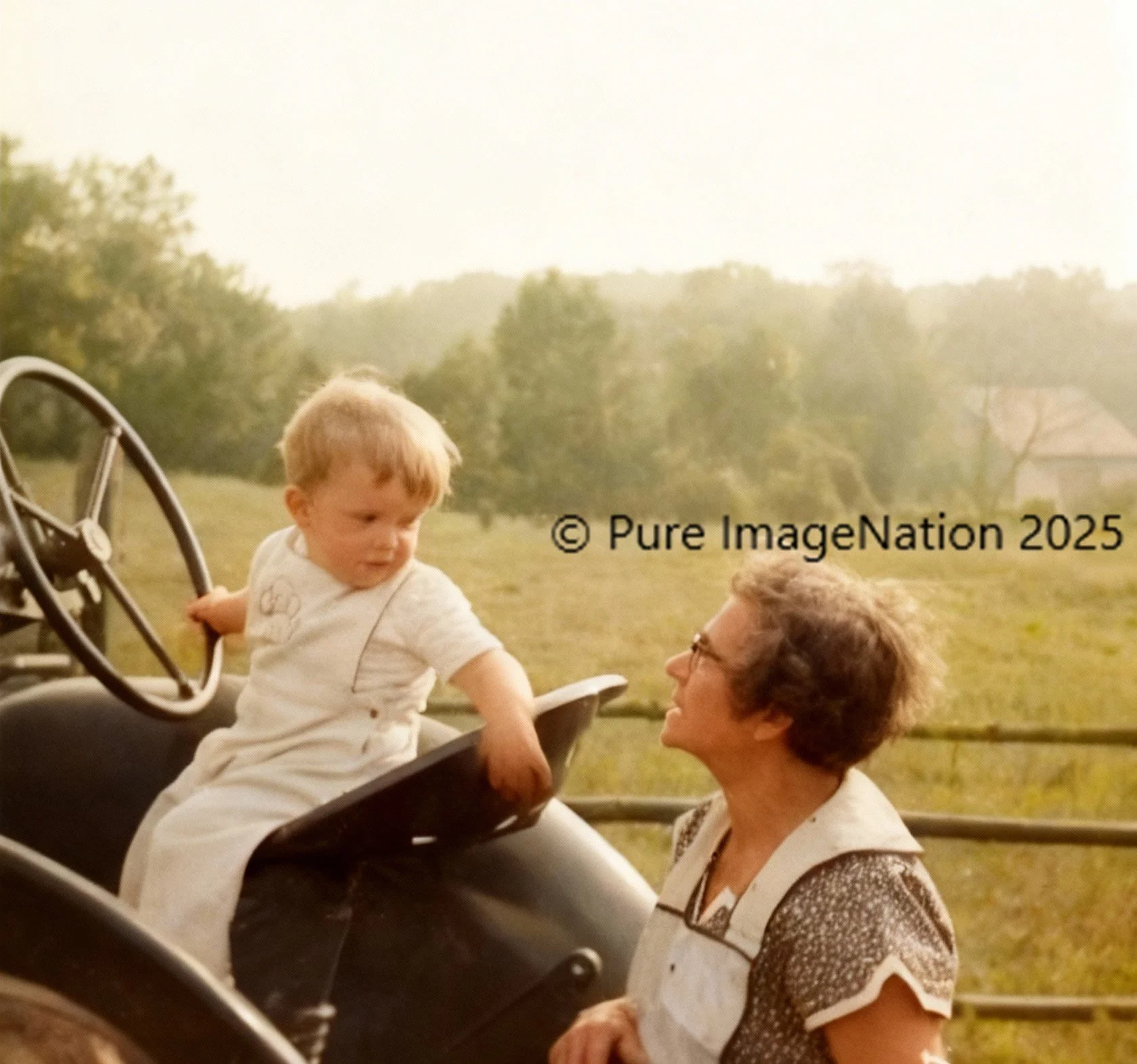 Child sitting on a tractor, looking at an adult, farm land in the background.