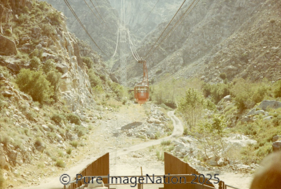 A cable car traveling across mountainous terrain with rocky slopes, sparse vegetation, and a dirt path below.