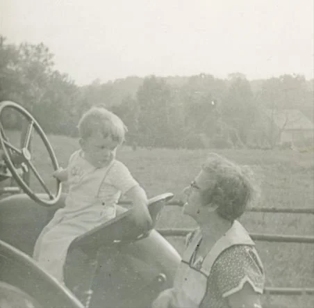 Child sitting on a tractor, looking at an adult, farm land in background