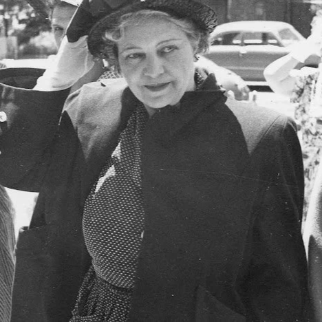 A woman dressed in a coat and dress from the 1950s stands with her hand on her hat looking to the right.