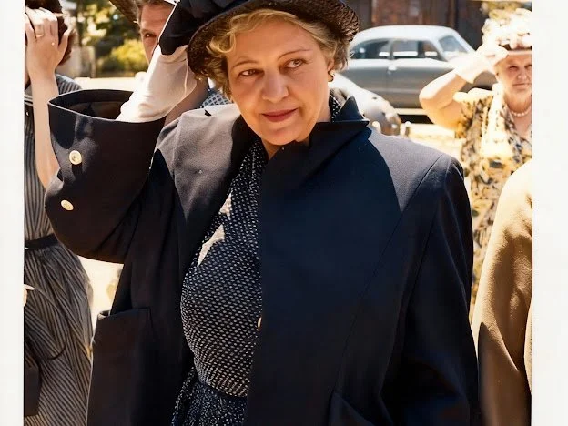 Color photo of a woman from the 1950s wearing a black coat and printed dress. Her hand is on her hat and she is looking to the right.