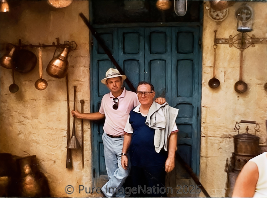Two men standing in front of a blue wooden door, surrounded by hanging copper pots and old kitchen utensils on a rustic wall.