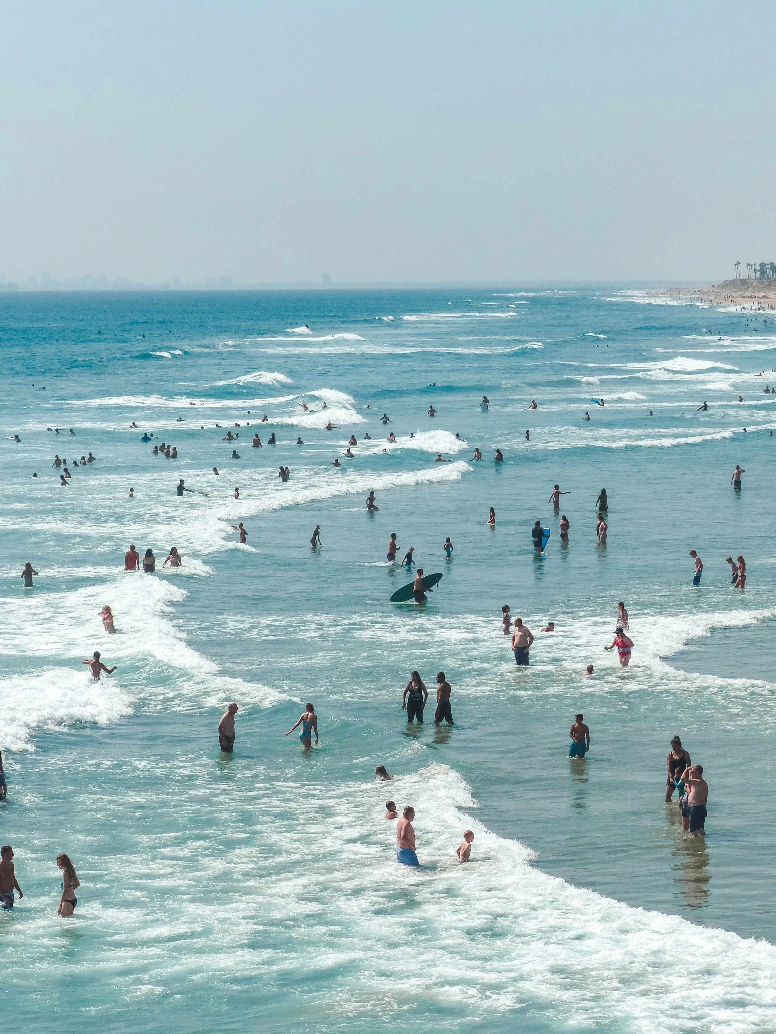 Crowded beach with numerous people swimming and playing in the ocean, waves approaching the shoreline under a clear sky.