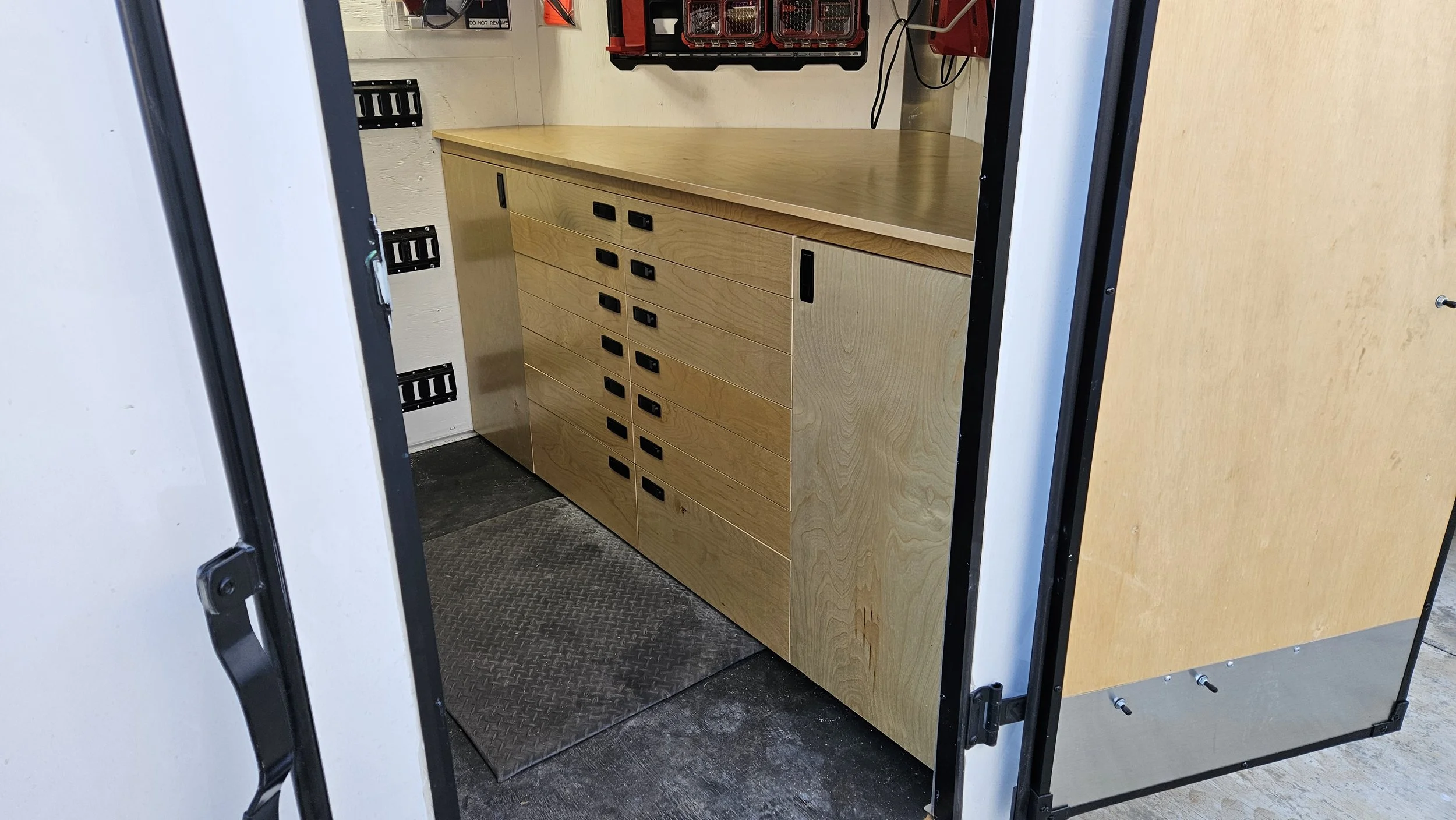 Inside of a mobile workshop or storage van showing a wooden cabinet with multiple drawers and a countertop, with electrical panels and wires on the wall above.
