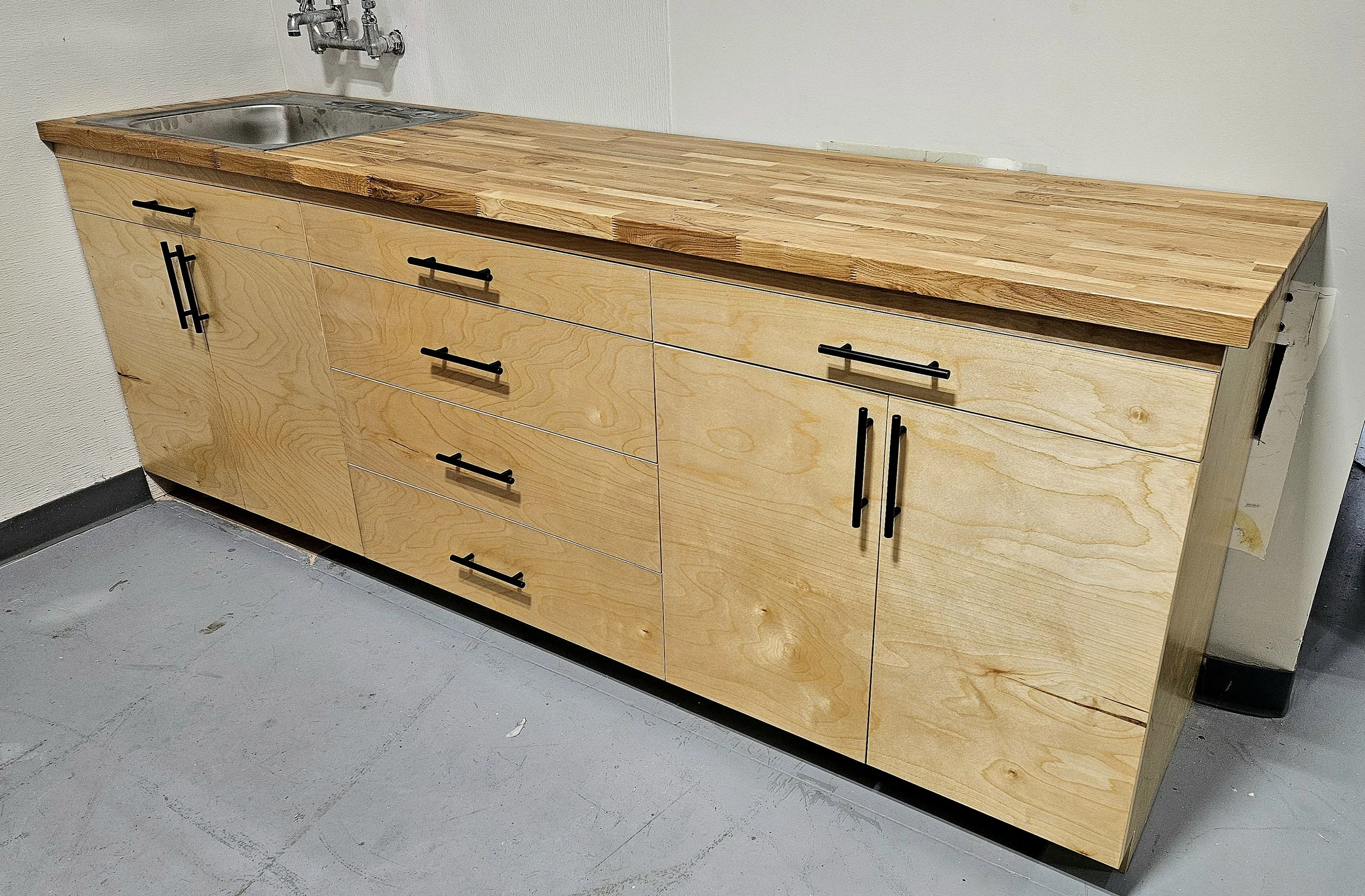 A wooden cabinet with multiple drawers and black handles is placed against a light-colored wall in a room with a concrete floor. The countertop is made of wood as well and has a small stainless steel sink with a wall-mounted faucet.