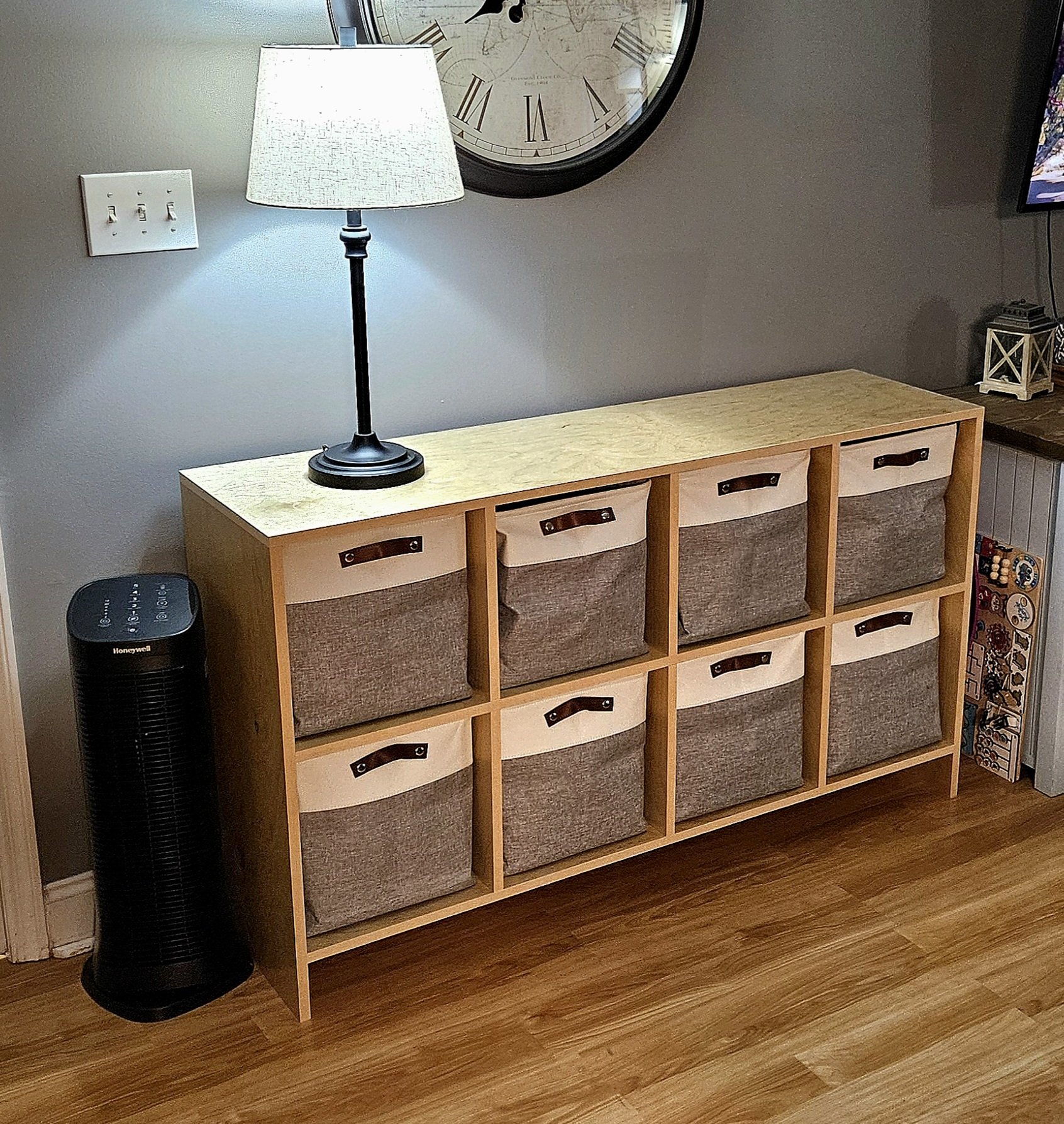 A wooden storage unit with nine fabric bins, a table lamp on top, a large round clock on the wall above, a black tower fan to the left, and a partial view of a TV and some books or games to the right.