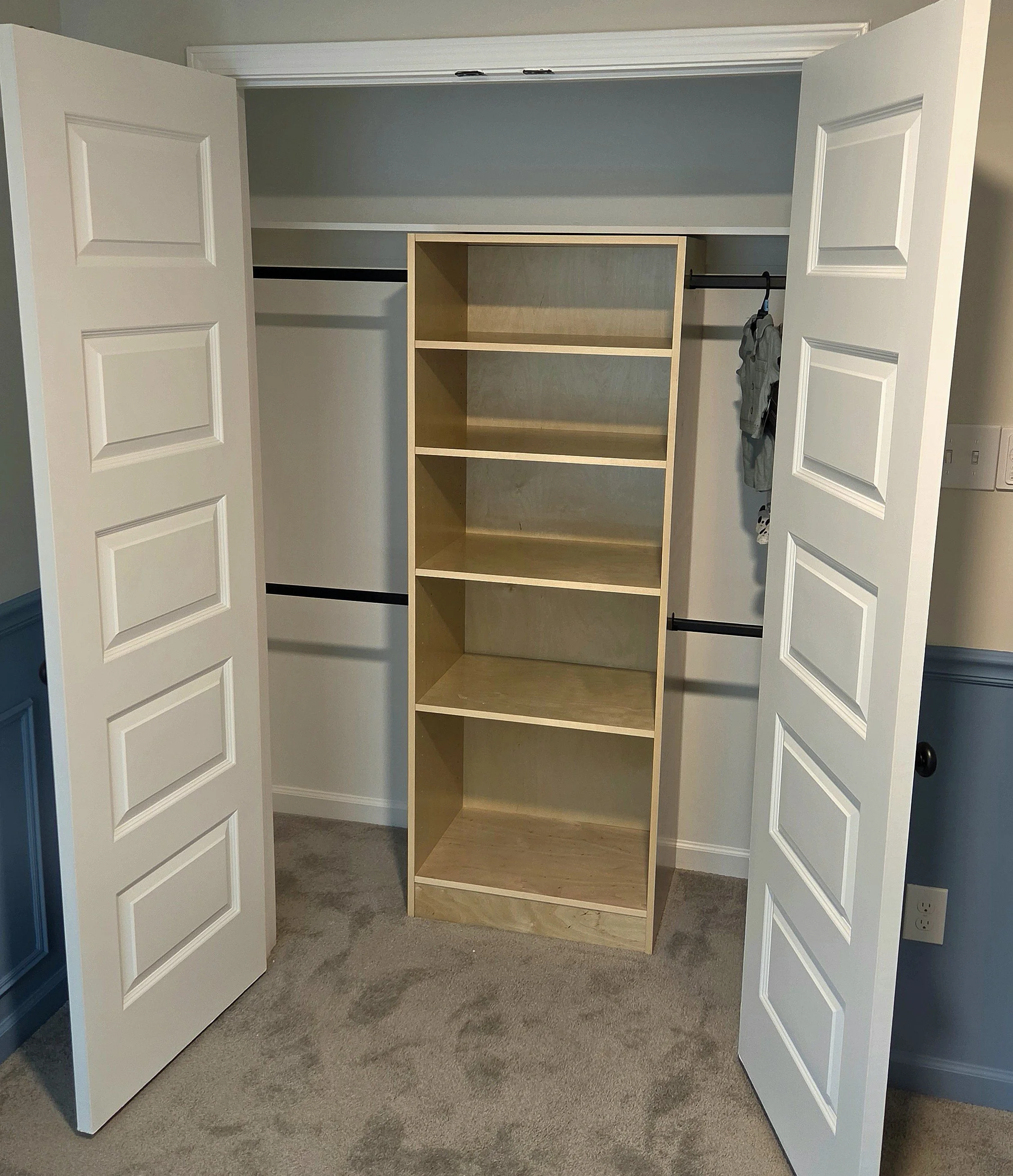 A closet with double white doors open, revealing an empty wooden shelving unit in the center. The closet has hanging rods on either side of the shelving, with some clothing items hanging on the right side. The floor is carpeted, and the walls are painted gray.