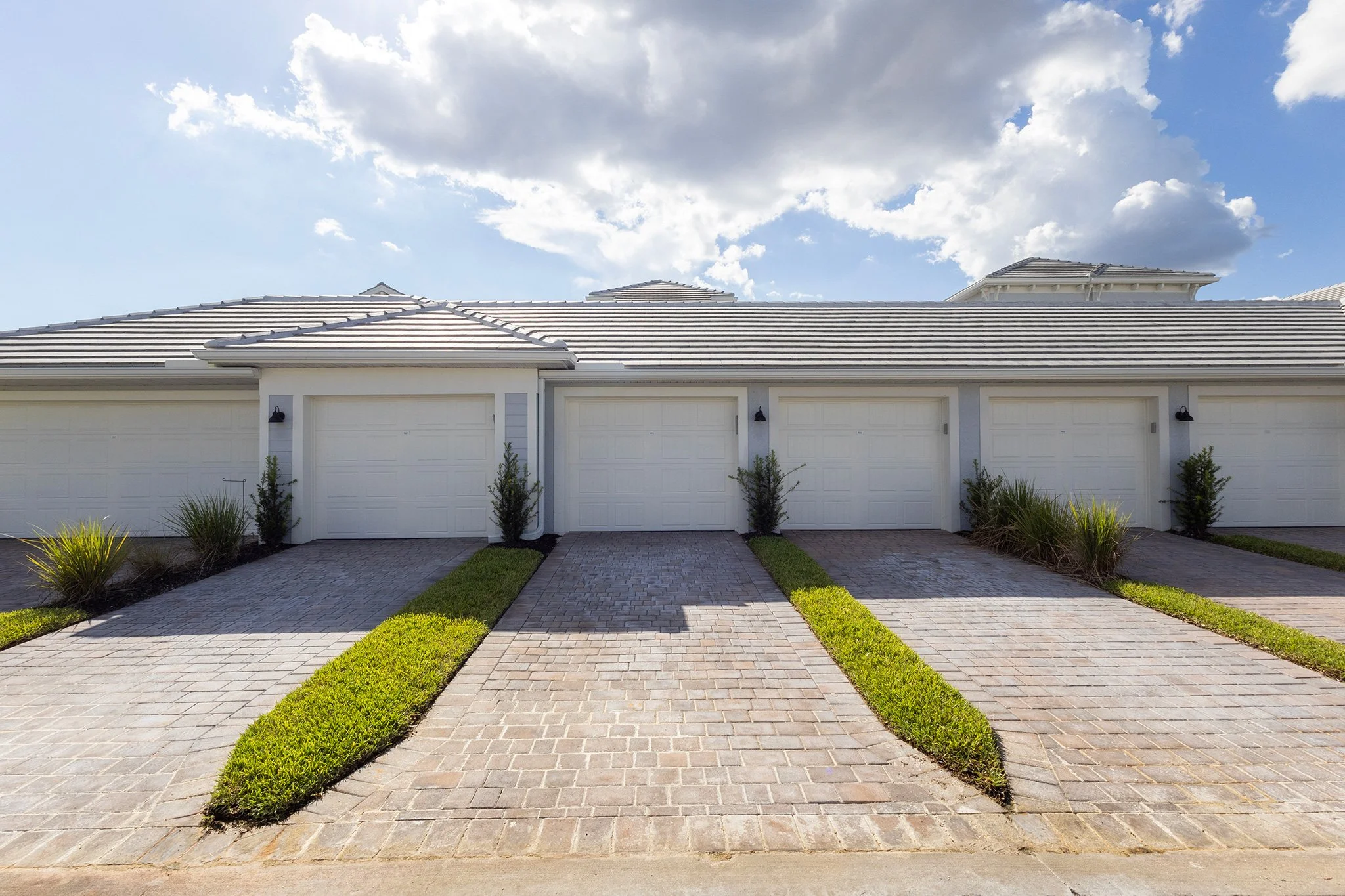 Row of white garage doors with small shrubs and greenery in front, gray brick driveways, and blue sky with clouds above.