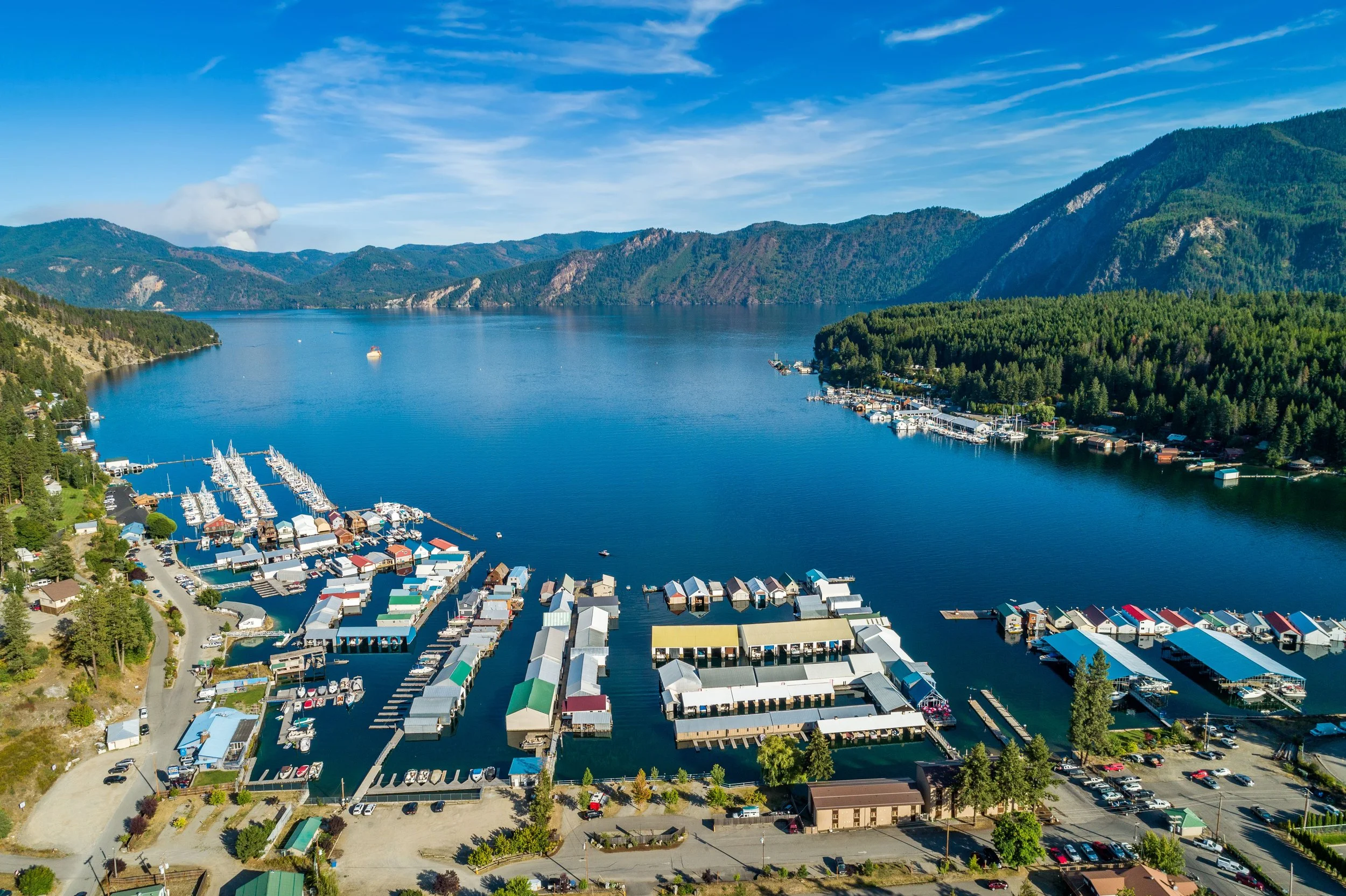 Aerial view of a marina with boats and docks on a large lake surrounded by mountains and lush forests under a blue sky with scattered clouds.