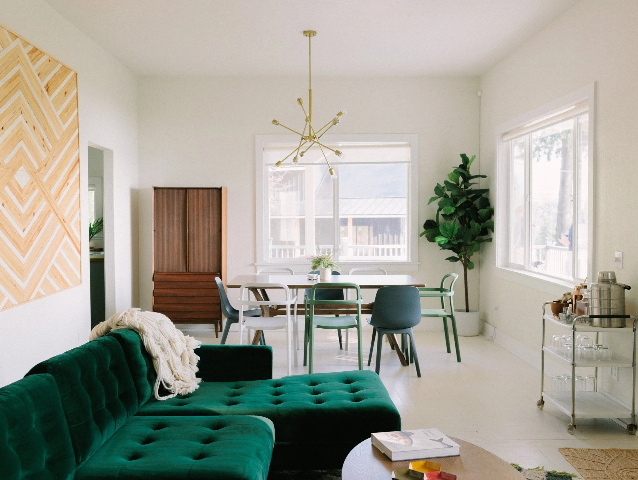 Bright dining and living room with large windows, green velvet sofa, modern chandelier, dining table with mixed chairs, large potted plant, wooden sideboard, and a metal cart with glasses and beverages.