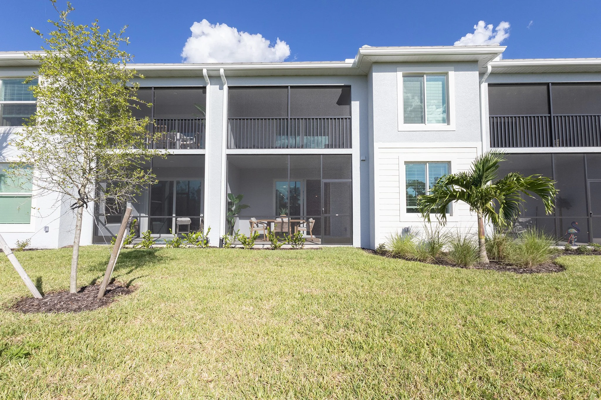 Exterior view of a modern apartment building with screened balconies, a lawn, a small tree, a palm tree, and landscaping under a blue sky with clouds.