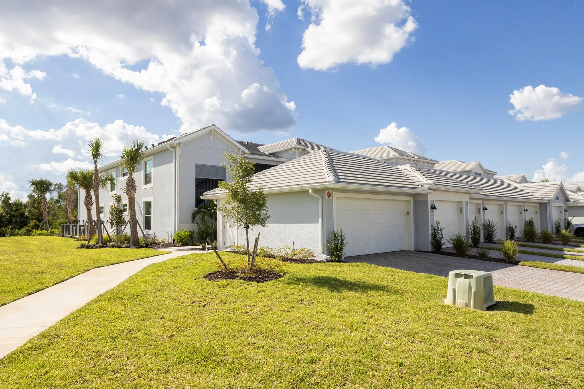 A white residential building with a tiled roof, attached garage, and a well-maintained lawn with young trees and palm trees, under a partly cloudy sky.