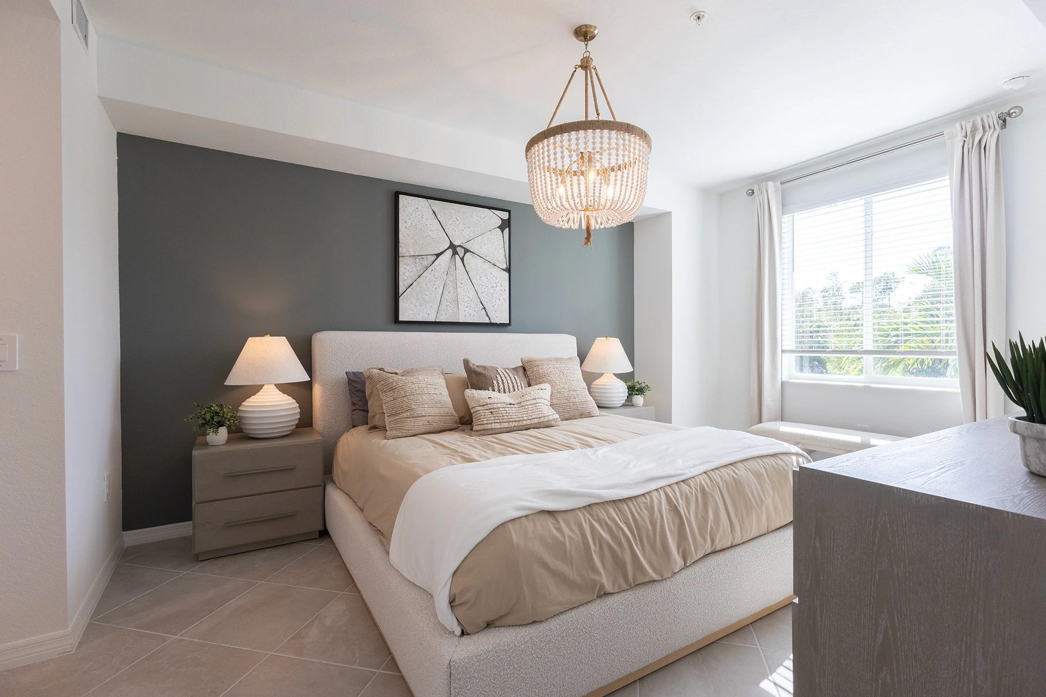 Bright bedroom with a large window, beige bed with pillows, two nightstands with lamps, and a chandelier hanging from the ceiling, gray and white walls.