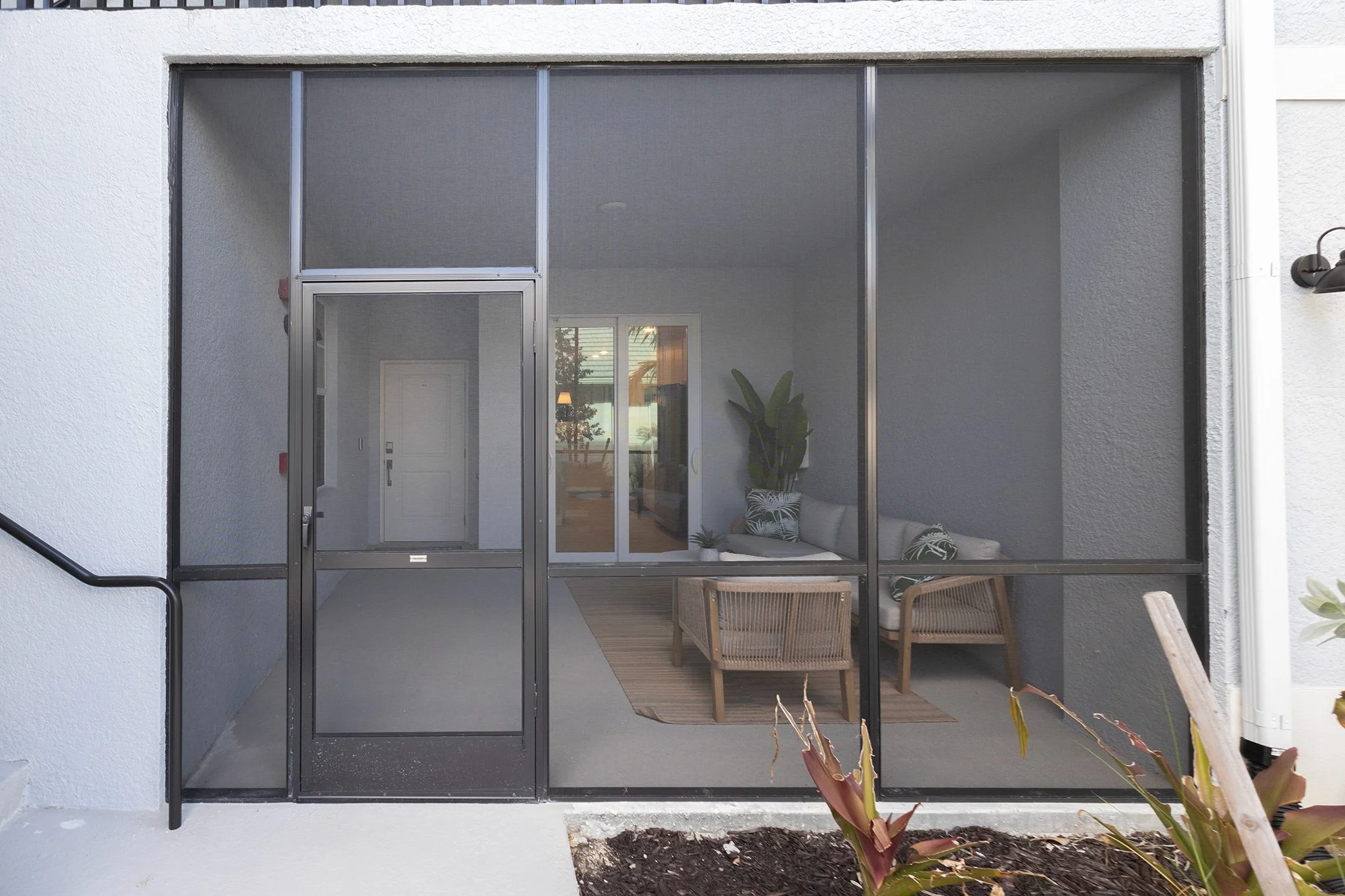 The image shows a screened-in porch or patio area with a glass sliding door leading to the interior of a house. Inside, there is a small seating area with a white sofa, a wooden armchair, and some decorative pillows. There are also indoor plants and