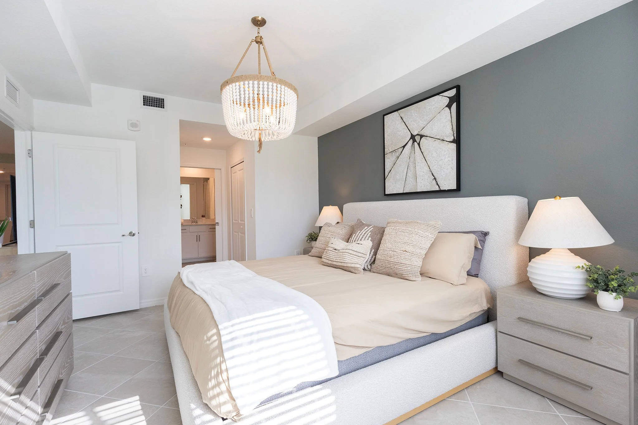A modern bedroom with a white bed and beige bedding, dark gray accent wall with abstract art, two nightstands with lamps, and a ceiling chandelier, with a view into an adjoining bathroom.