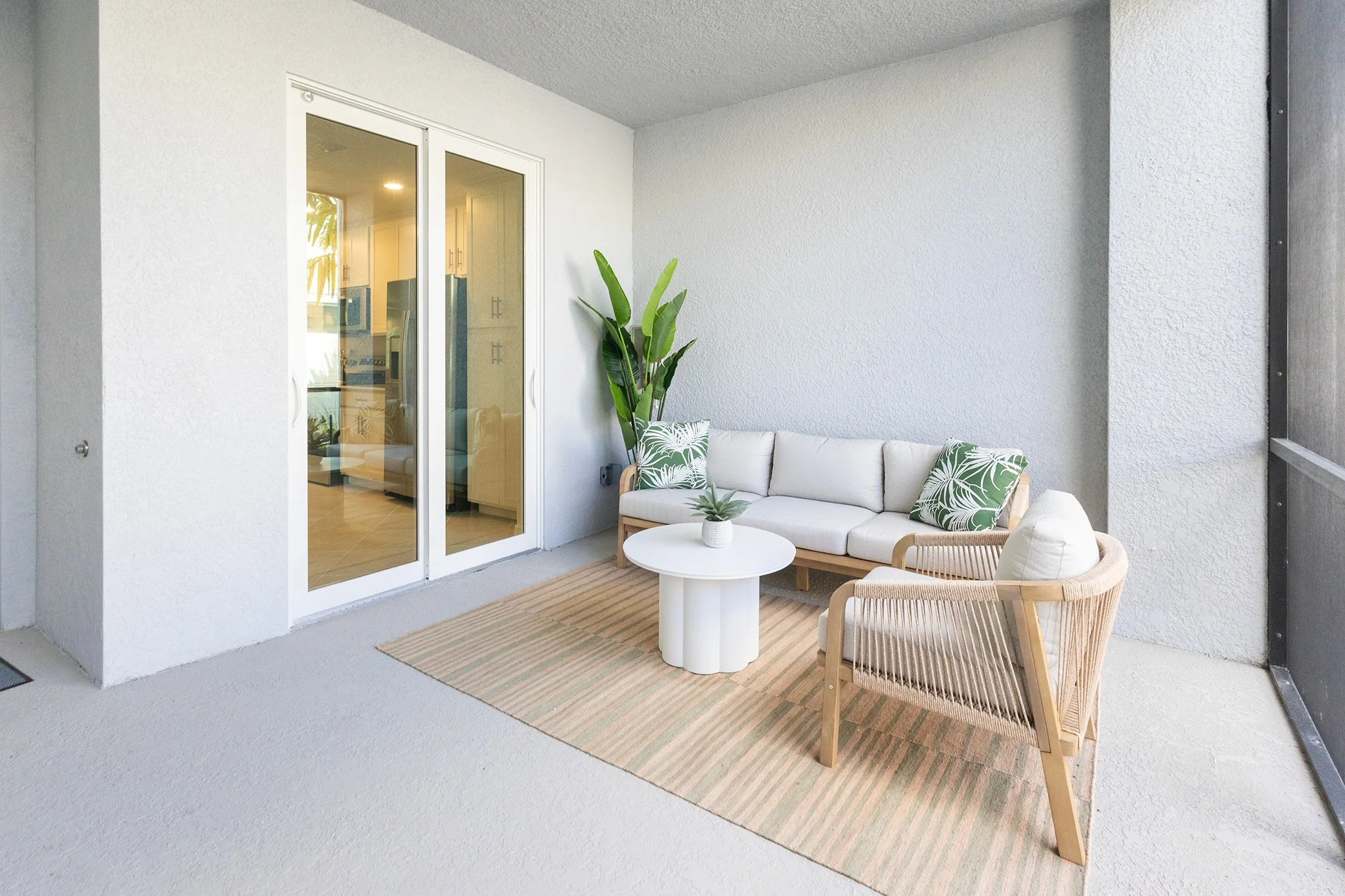 Patio with white couch, wicker armchair, round white coffee table, potted plant, large windowed doors, and a potted plant in the background