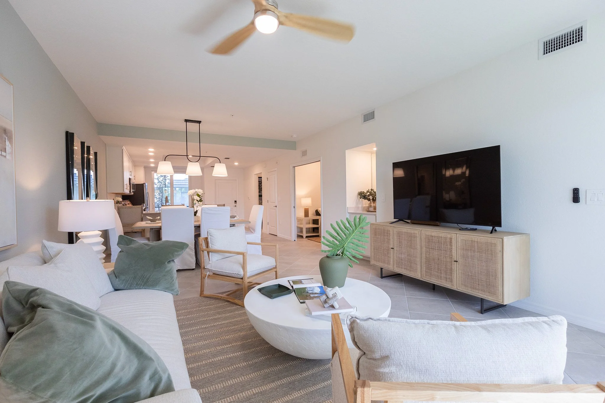 Bright open-concept living room and dining area with neutral tones, a wall-mounted flat-screen TV on a rattan cabinet, a white sofa with green and cream pillows, a white round coffee table with magazines, a green plant in a vase, and a dining table w