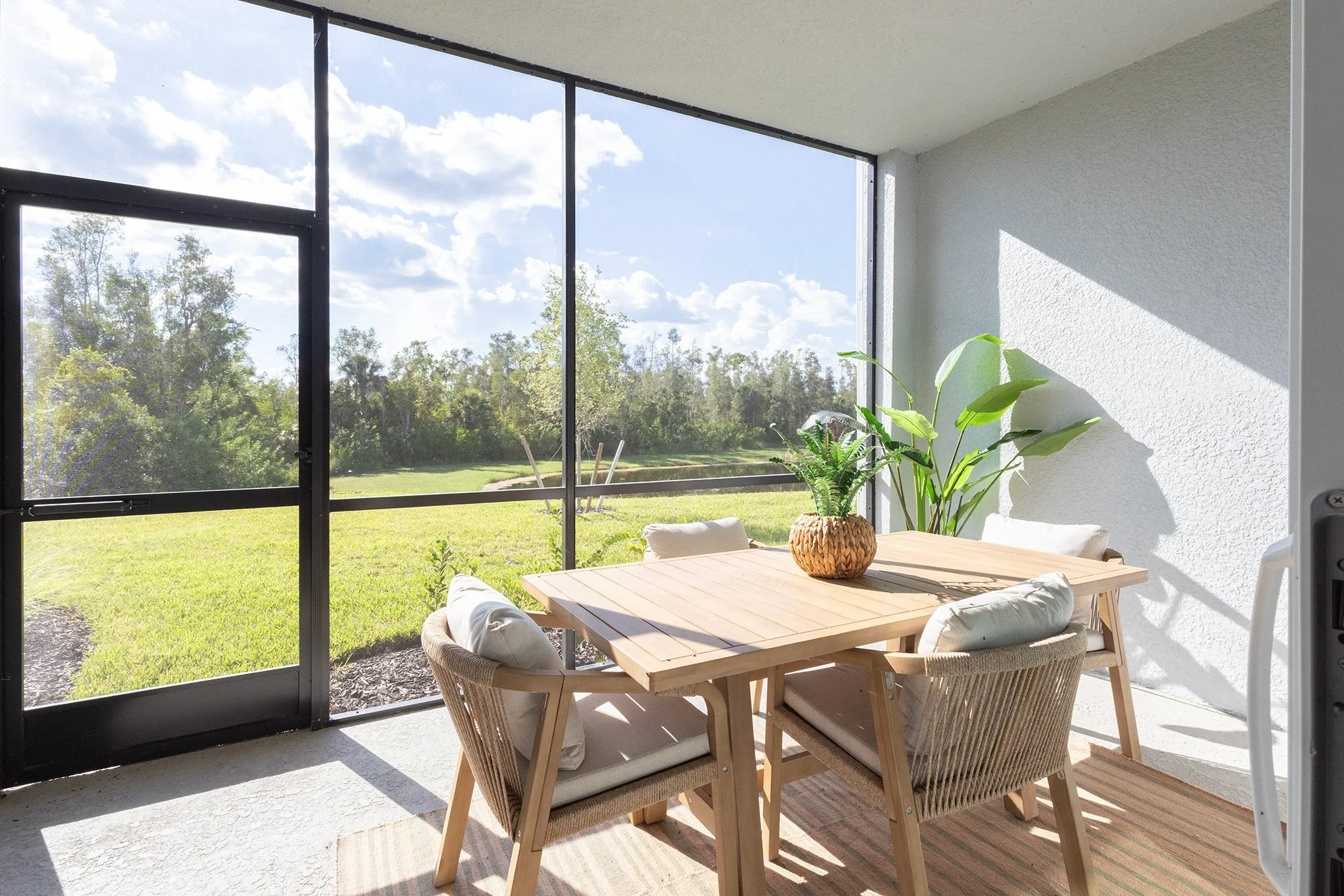 Indoor patio with a wooden dining table, four cushioned chairs, a potted plant, and sunlight streaming through large glass windows overlooking a green lawn and trees.
