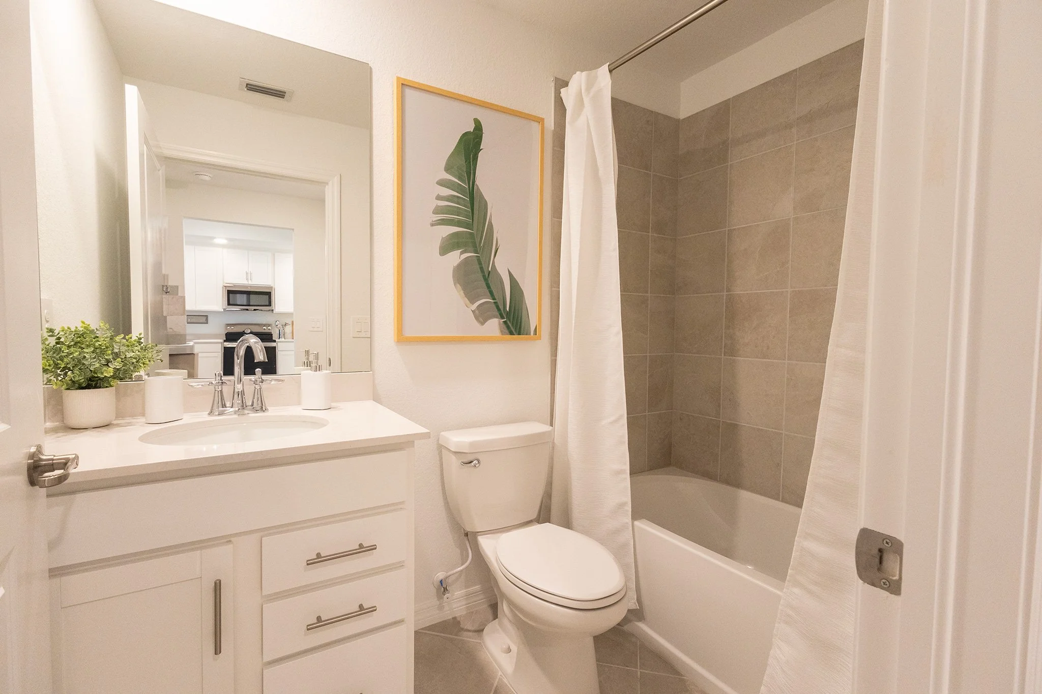 A modern bathroom featuring a white vanity with a sink, a large mirror, potted plant, framed leaf artwork on the wall, a toilet, and a bathtub with beige tile surround and a white shower curtain.