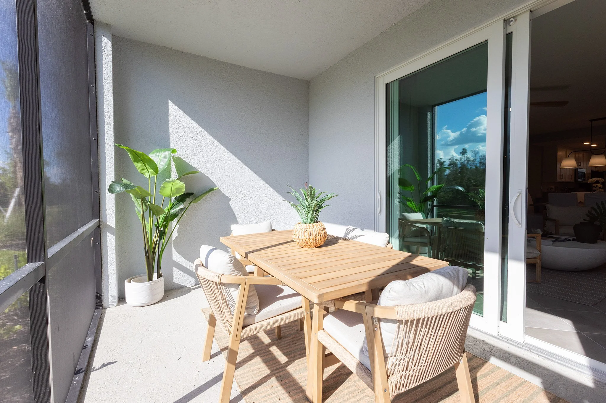 Balcony with a wooden table, four chairs with cushions, large potted plant, and glass sliding door reflecting the blue sky and clouds.