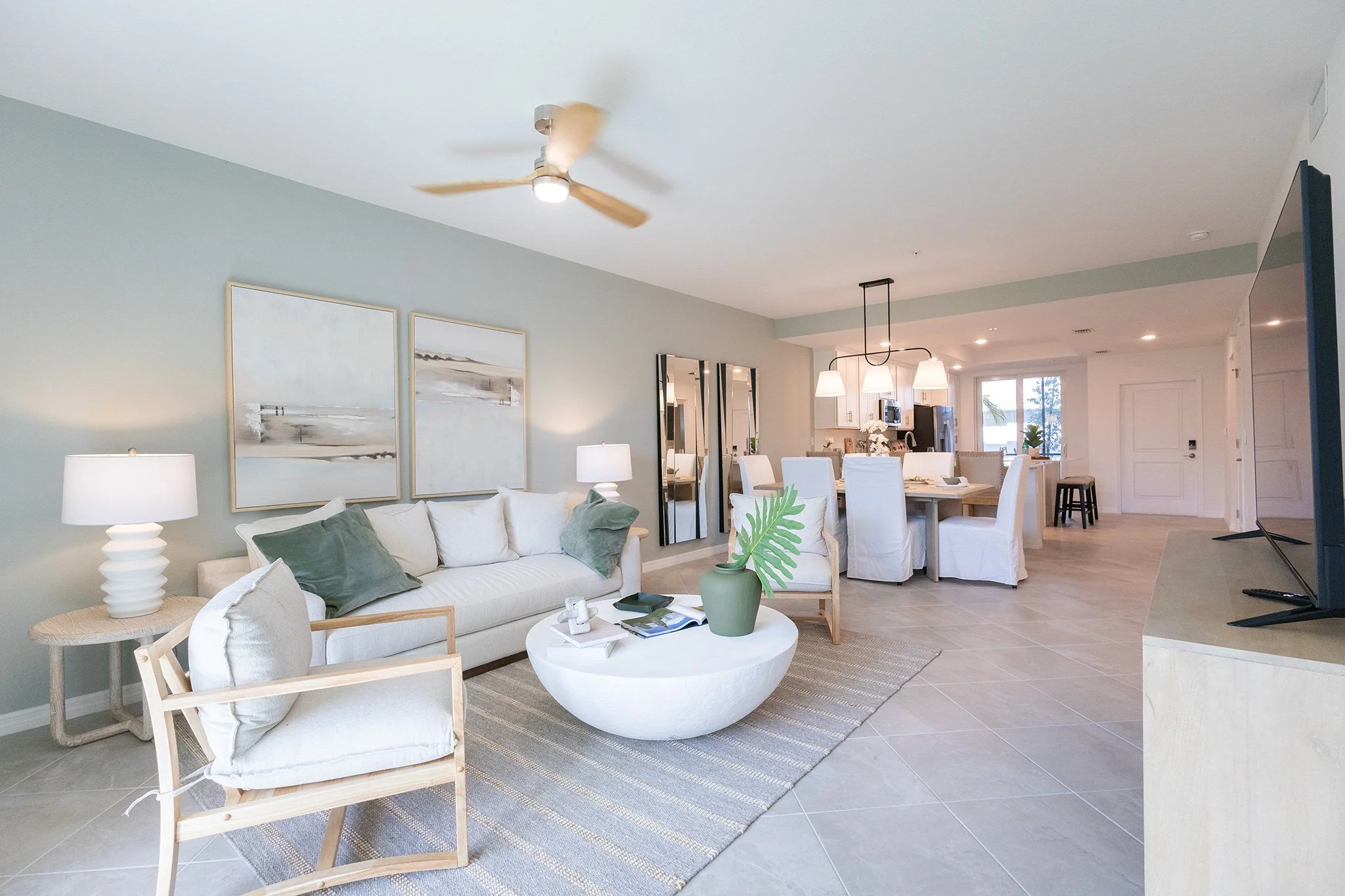 Living room with a white sofa, armchair, coffee table with green vase, lamps, and wall art, leading to dining area with white chairs and pendant lights, open view to kitchen in the background.