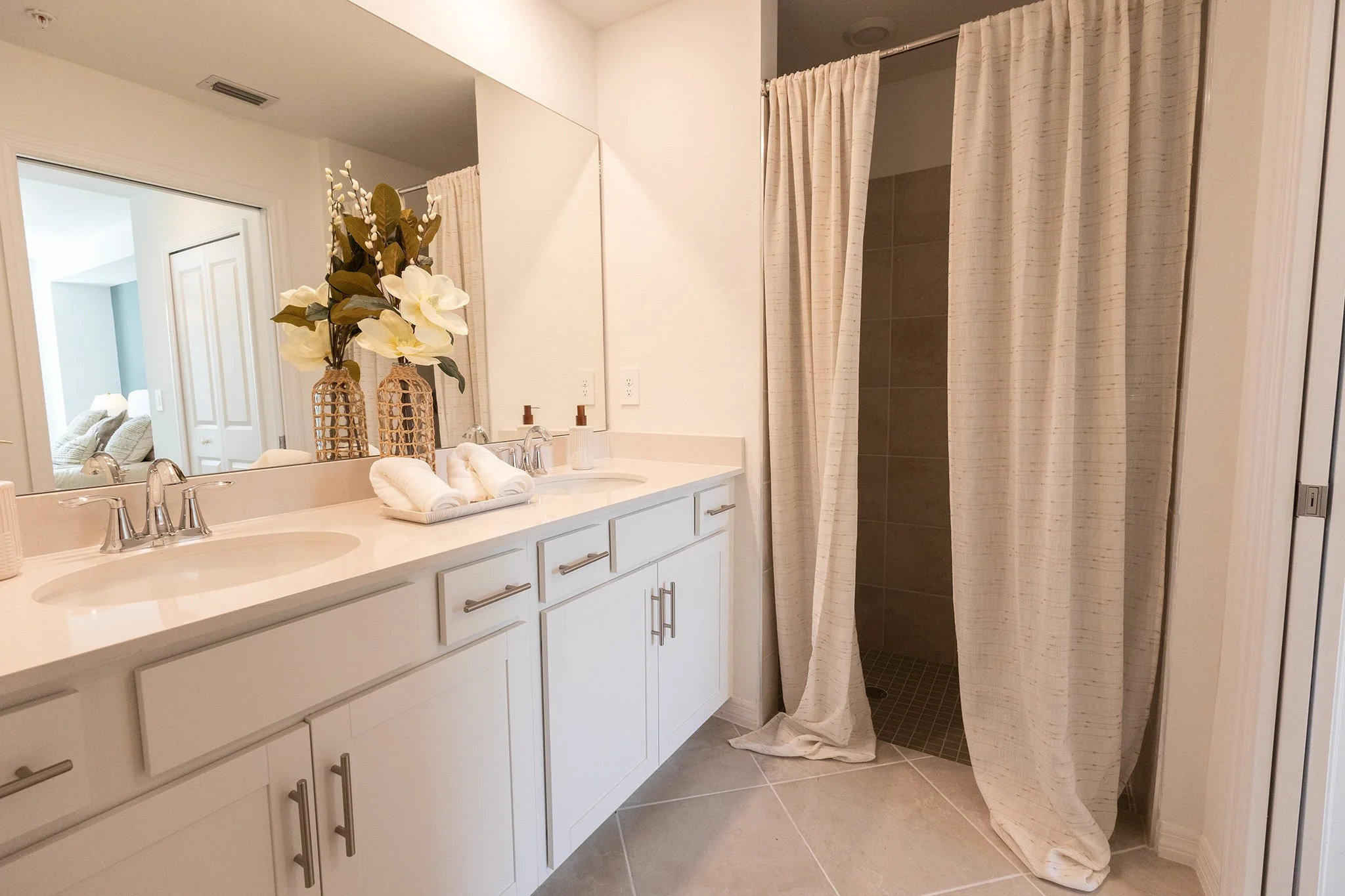 Bathroom with a double vanity, large mirror, and two vases with flowers. There are rolled towels on the counter. To the right, a shower with a beige curtain, beige tile walls, and a tiled floor.