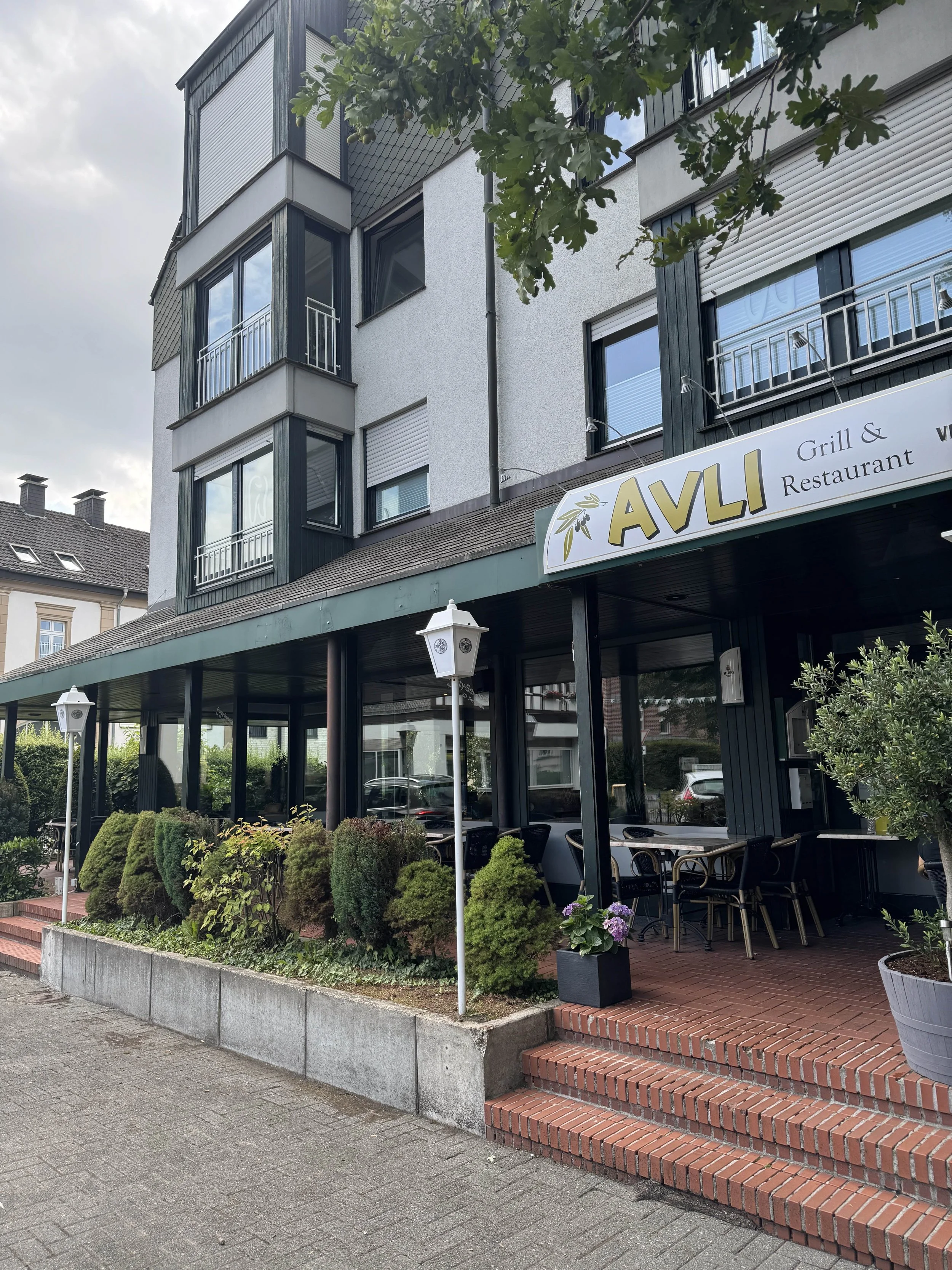 Exterior of a building with a restaurant named AVLI Grill & Restaurant, featuring outdoor seating and landscaped bushes.