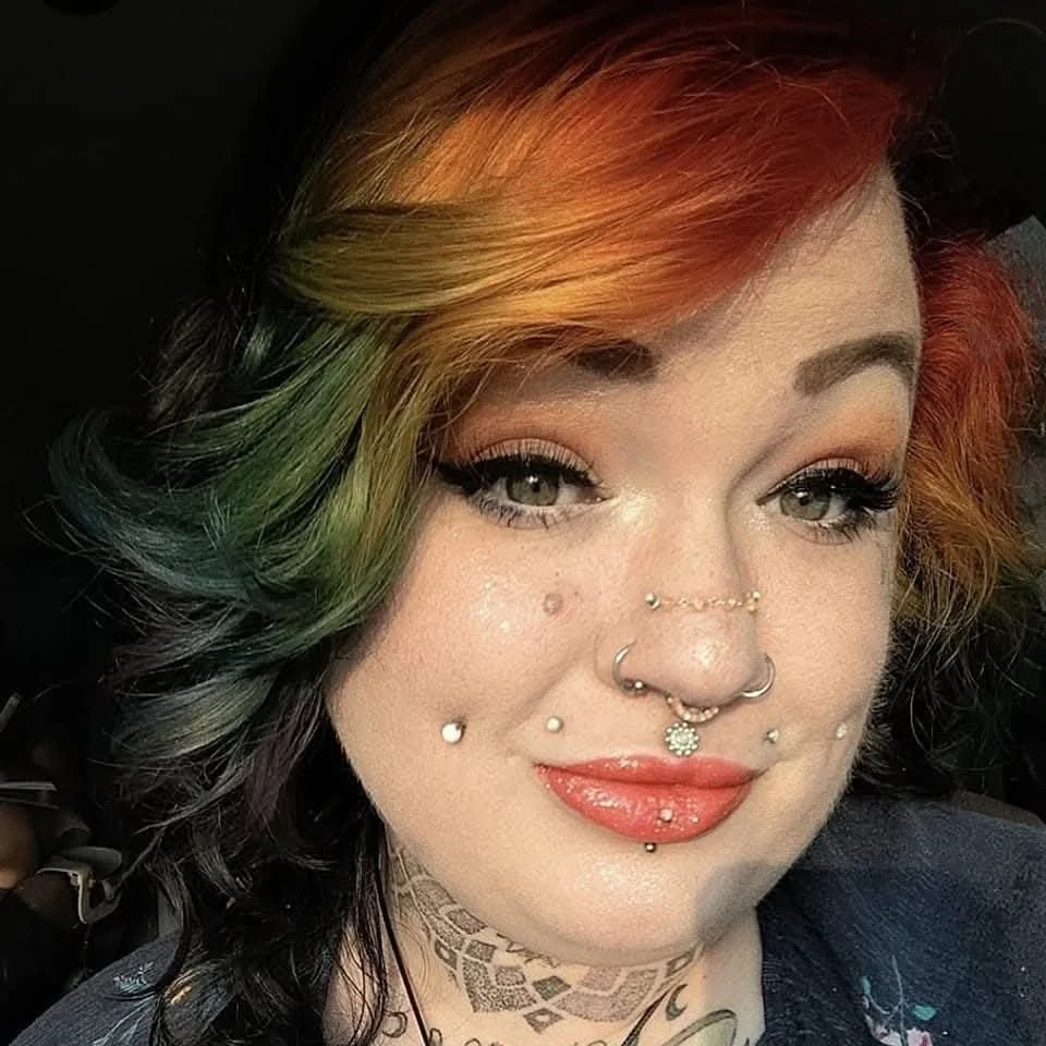A woman with multicolored hair, featuring shades of orange, green, and black, with several facial piercings, including a septum, nostrils, lips, and cheeks, and tattoos on her neck.