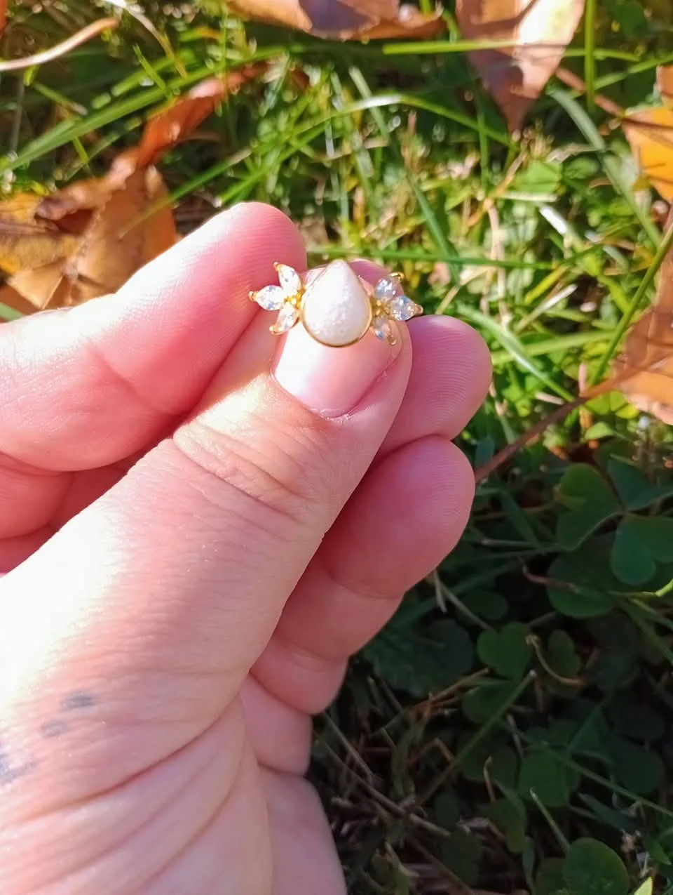 A hand holding a ring with a white teardrop-shaped gemstone surrounded by small clear stones, set in a gold band, in an outdoor green leafy setting.